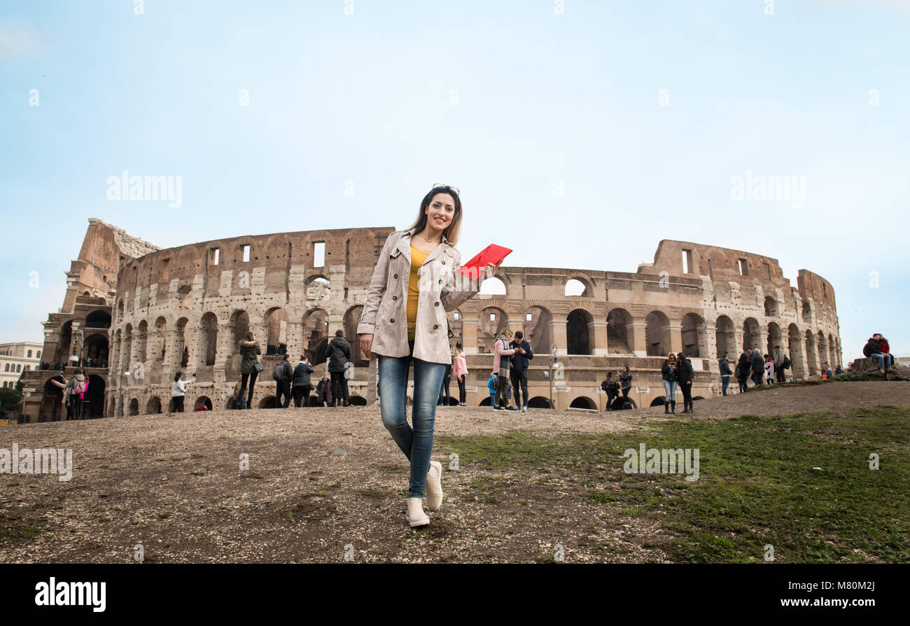 Junge hübsche touristische Frau mit roten Reisen buchen Sie lächelnd auf Kamera, im Hintergrund Kolosseum Monument in Rom Italien Stockfoto