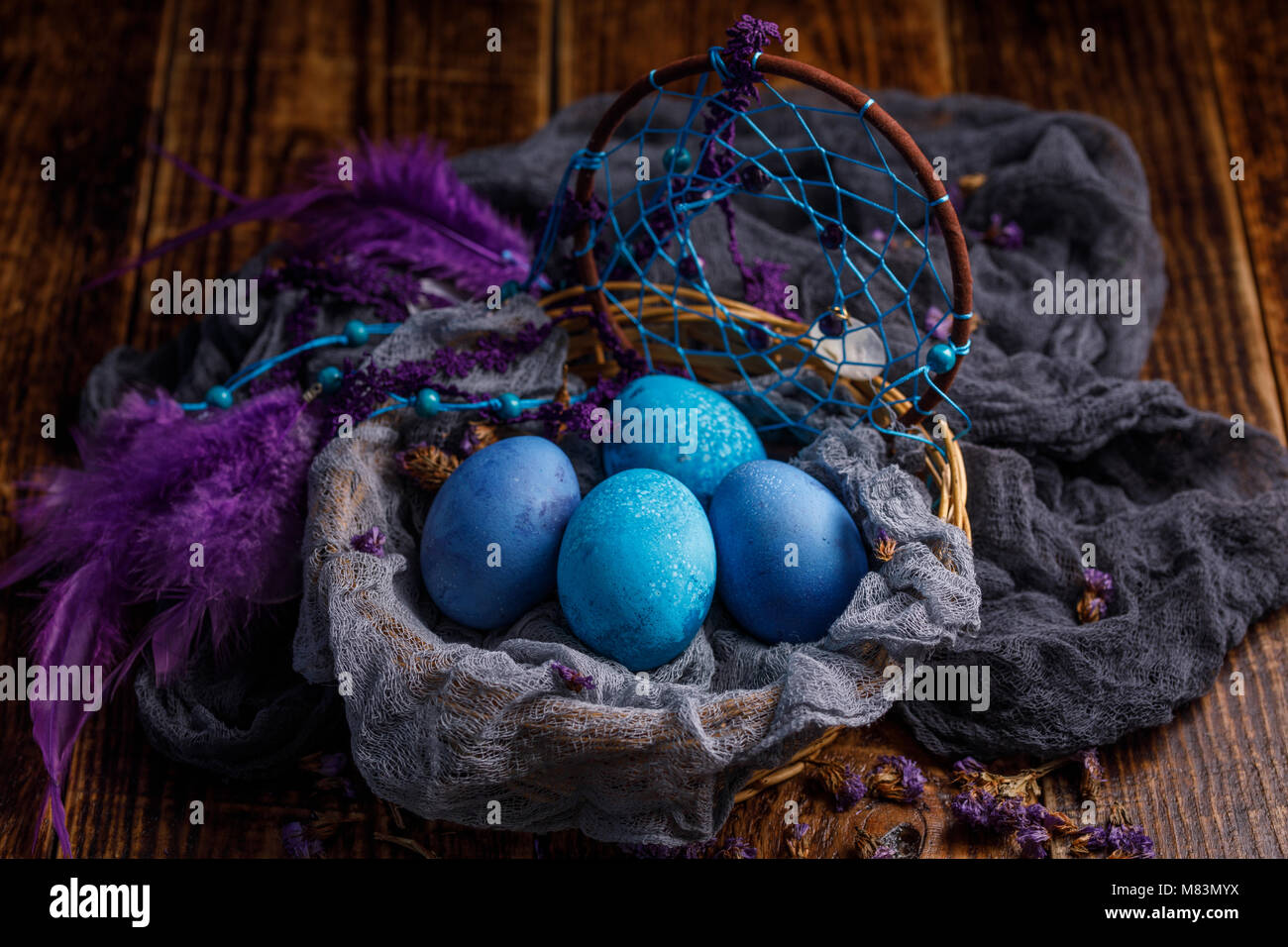 In Dunkelblau und blau lackiert, Eier in einem Weidenkorb wie ein Nest mit Federn aus einem Catcher Träume eingerichtet. Still-life auf einem hölzernen Hintergrund. Stockfoto