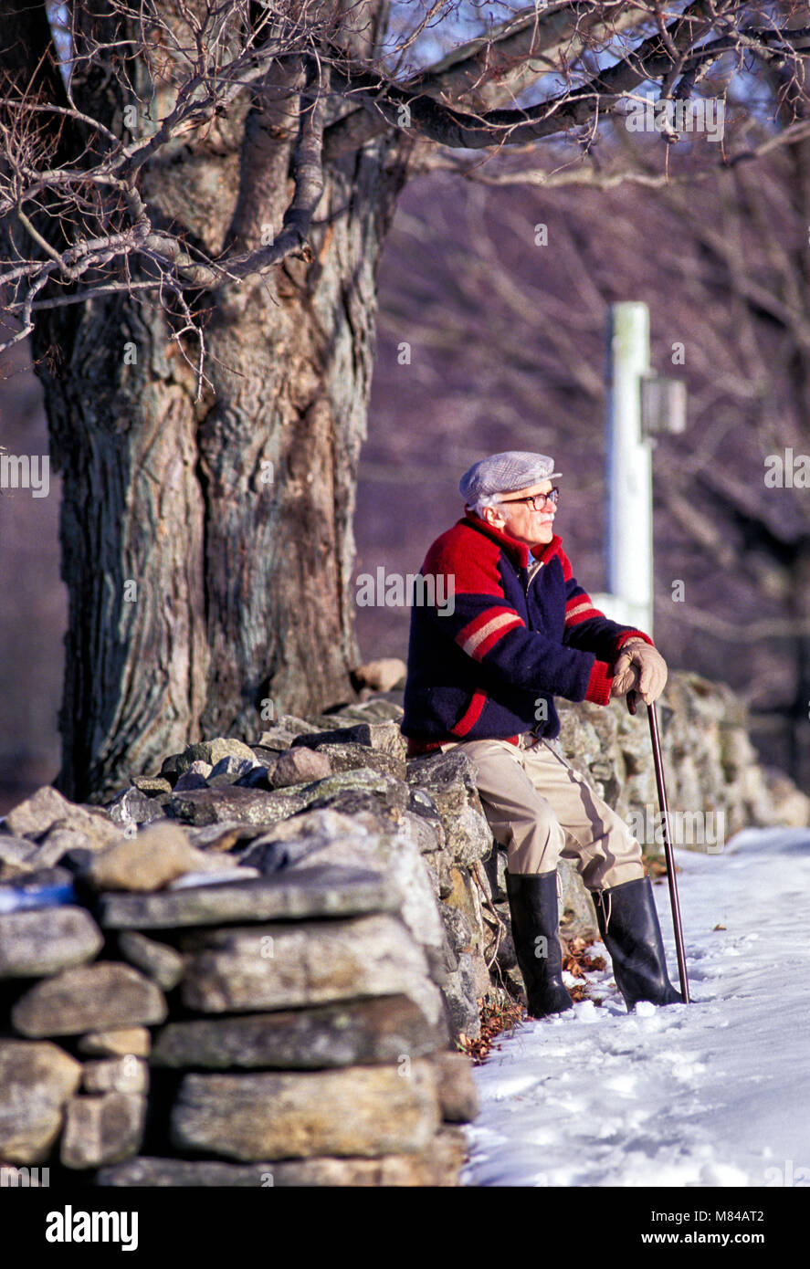 Älterer Mann sitzt allein an einem New England fieldstone Zaun an einem verschneiten Tag Stockfoto