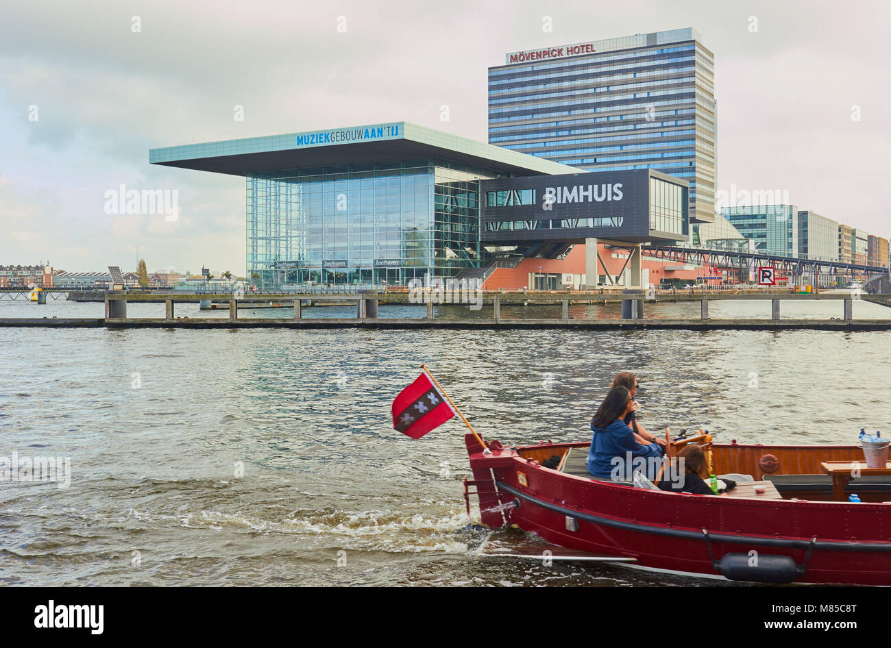 Boot mit Amsterdam Flagge das Bestehen der Bimhuis Konzerthalle, Teil der Muziekgebouw Komplex auf der IJ Fluss, Amsterdam, Niederlande. Stockfoto