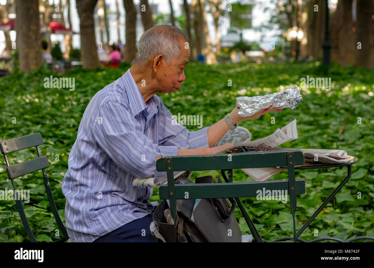 Ein chinesischer Mann sitzen an einem Tisch eine Zeitung lesen im Bryant Park, Manhattan, New York City Stockfoto