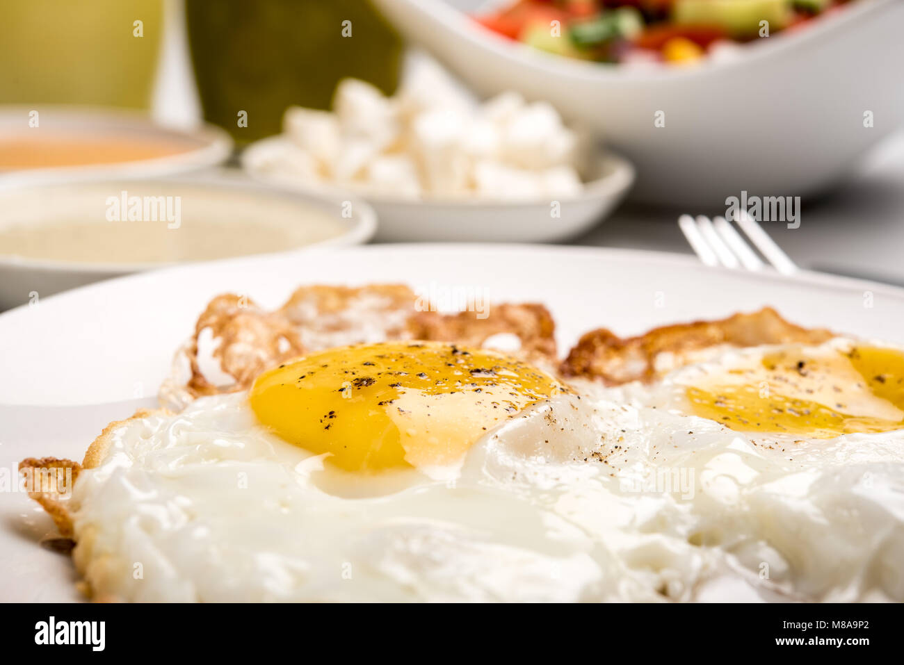 Traditionelles israelisches Frühstück mit zwei Spiegeleier, gelben Käse, Salat, eine frische Brötchen und einer Tasse Cappuccino. Nahaufnahme auf die gebratenen Eier Stockfoto