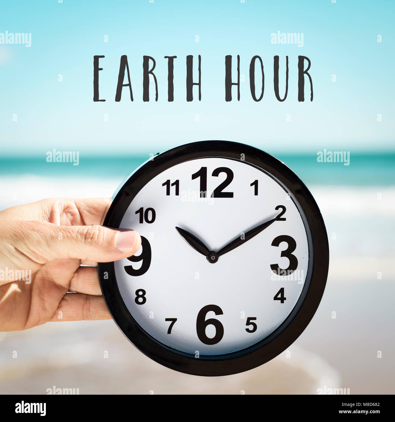 Nahaufnahme eines jungen kaukasischen Mann hält eine Uhr auf dem Strand vor dem Meer und der Text der Earth Hour Stockfoto