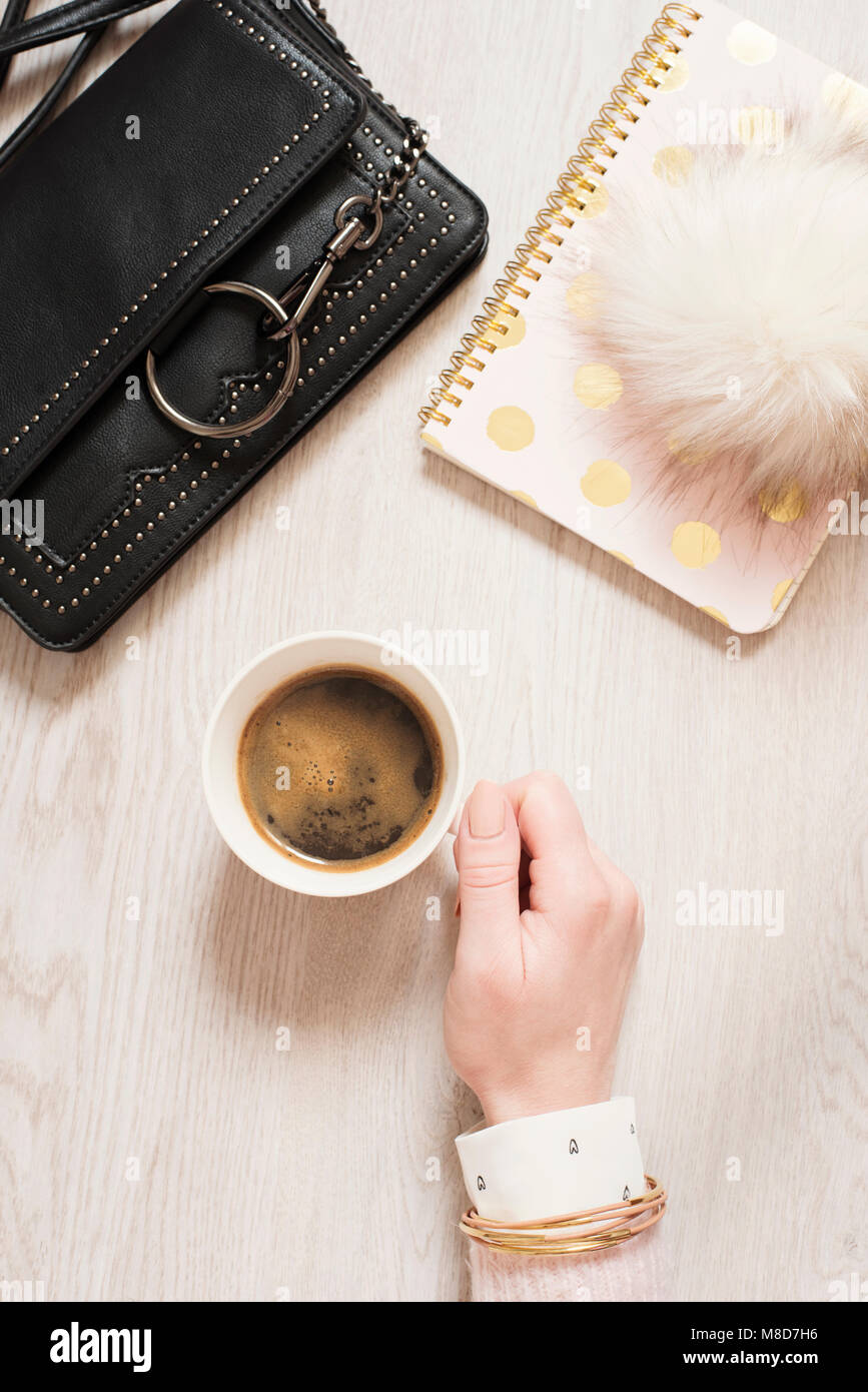 Ansicht von oben Frau Hand, die eine Tasse schwarzen Kaffee auf Flach. Arbeitsbereich mit Notebook in Pink und Gold, Mode Handtasche und Fell Kugel Stockfoto