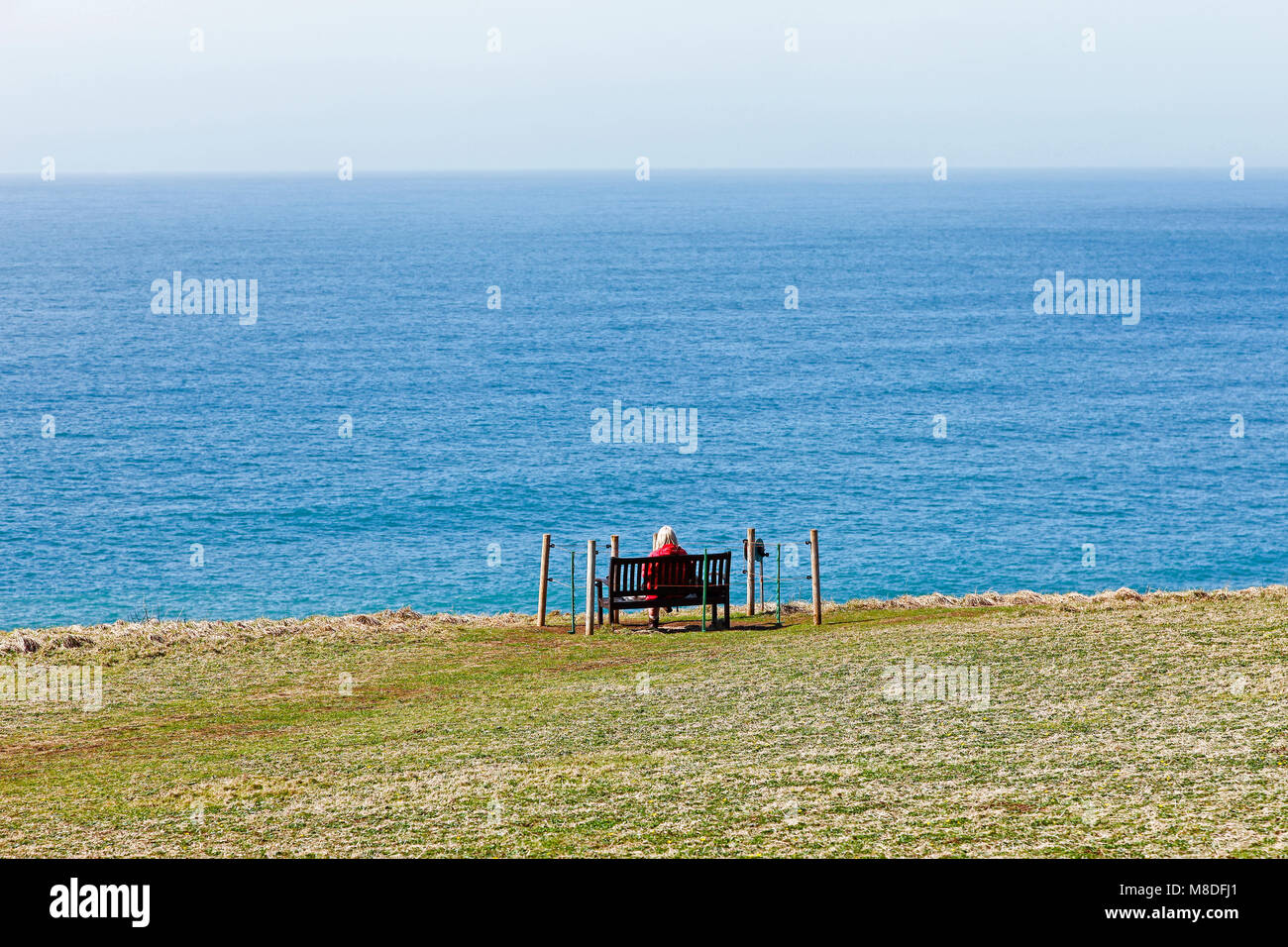 Eine Frau sitzt auf einem isolierten Sitzbank mit Blick aufs Meer auf der Lizard Halbinsel, Cornwall, South West England, Großbritannien Stockfoto