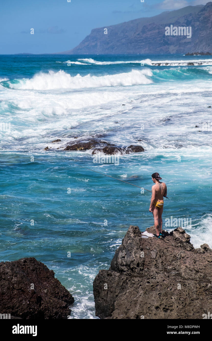 Jugend Angeln aus Felsen Tragen Badehose, Hut und Schuhe, Alcala, mit der Los Gigantes Felsen im Hintergrund, Teneriffa, Kanarische Inseln, Spanien Stockfoto