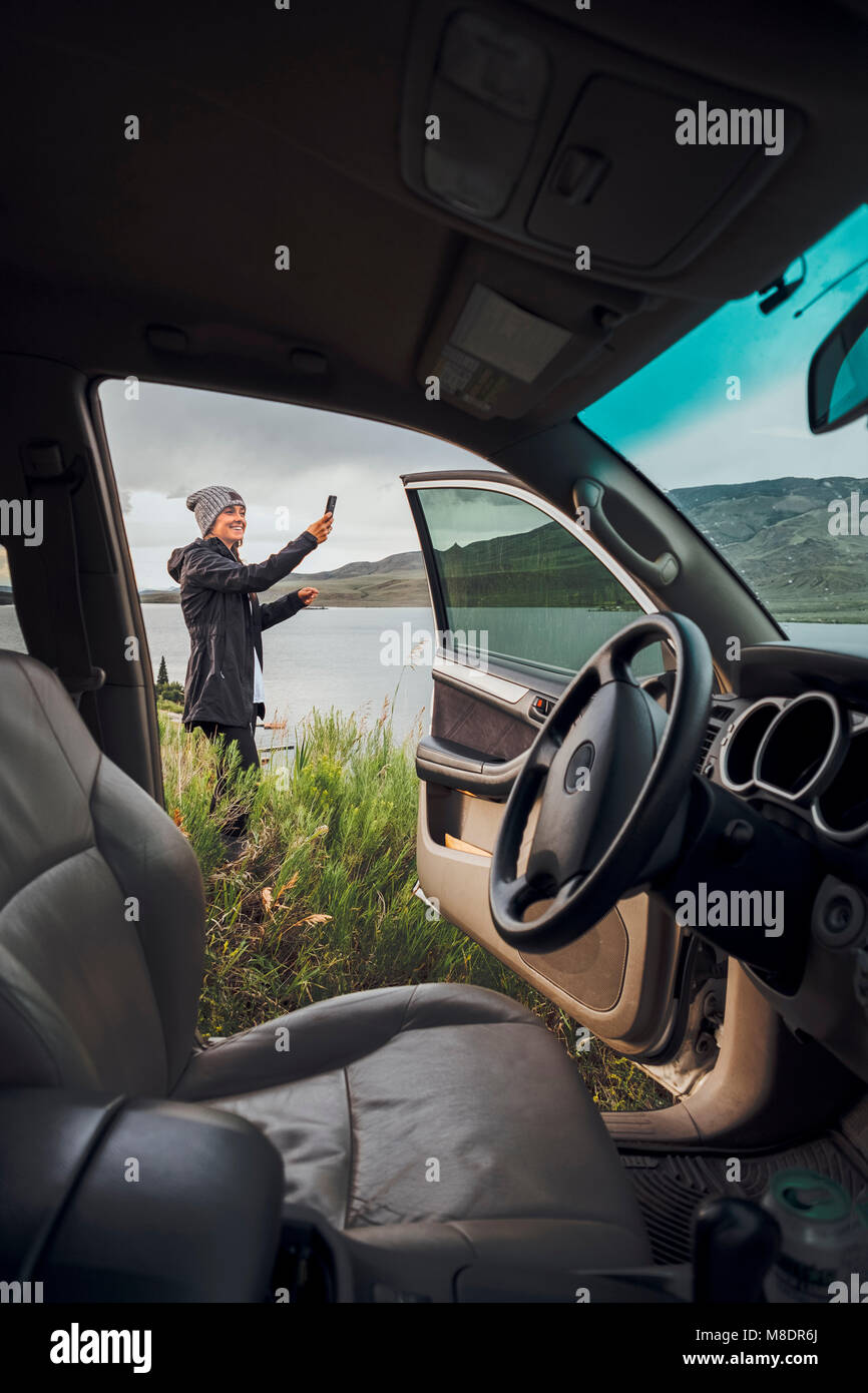 Junge Frau stand neben Dillon Reservoir, Smartphone, Blick durch die geparkten Auto, Silverthorne, Colorado, USA Stockfoto