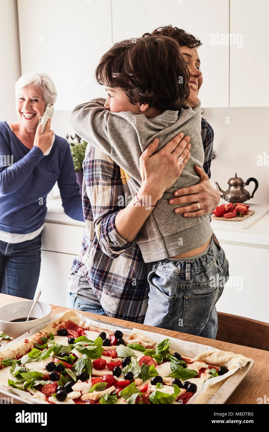 Mutter und Sohn umarmen in Küche, Großmutter im Hintergrund über Smartphone Stockfoto