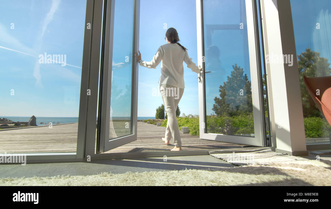 Schuss eine schöne Brünette mit losen Haare Öffnen von Türen auf den Balkon und Gehen auf die sonnige Terrasse mit Meerblick. Stockfoto