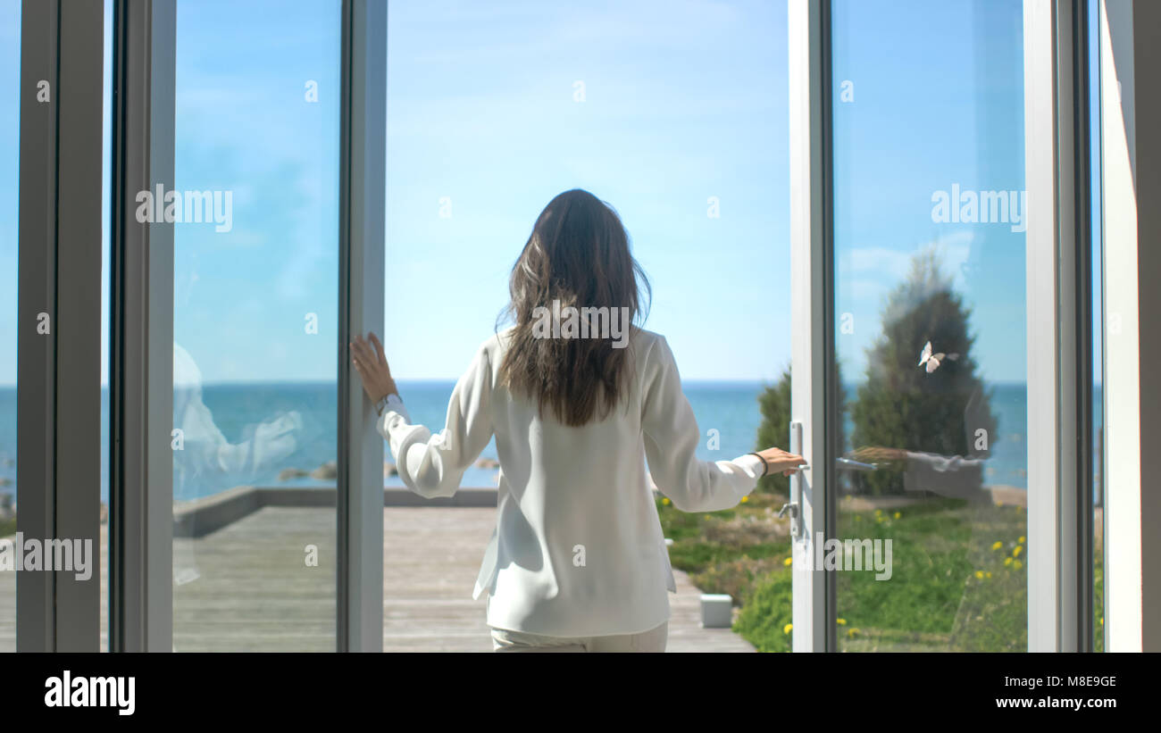 Schuss eine schöne Brünette mit losen Haare Öffnen von Türen auf den Balkon und Gehen auf die sonnige Terrasse mit Meerblick. Stockfoto