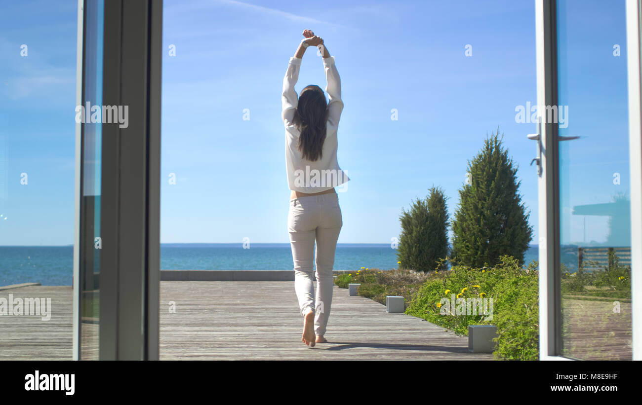 Schuss eine schöne Brünette mit losen Haare Öffnen von Türen auf den Balkon und Gehen auf die sonnige Terrasse mit Meerblick. Stockfoto