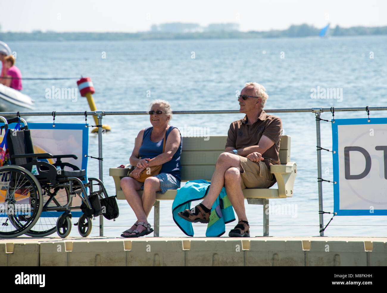 Älteres Paar entspannen Sie an der Wasserlinie auf einer Bank sitzen mit dem Rollstuhl für Neben der Bank gesperrt, am Haringvliet See in Hllevoetsuis Stockfoto