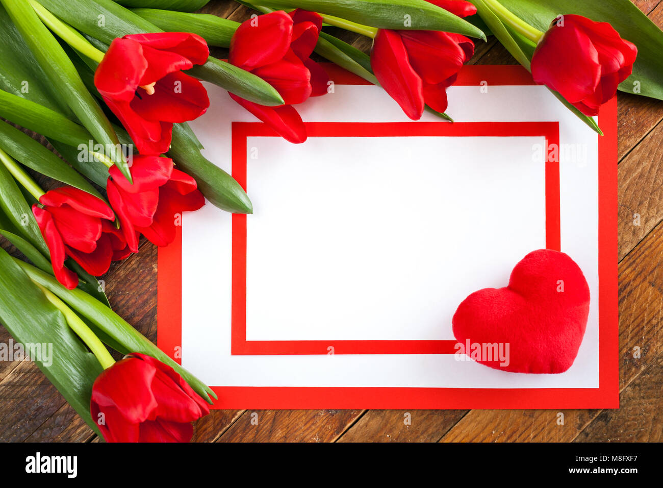 Grußkarte. Weißes leeres Blatt mit Plüsch Herz auf der Ecke, zwei roten Rahmen und rote Tulpen auf rustikalen diagonal Holz- Hintergrund. Stockfoto