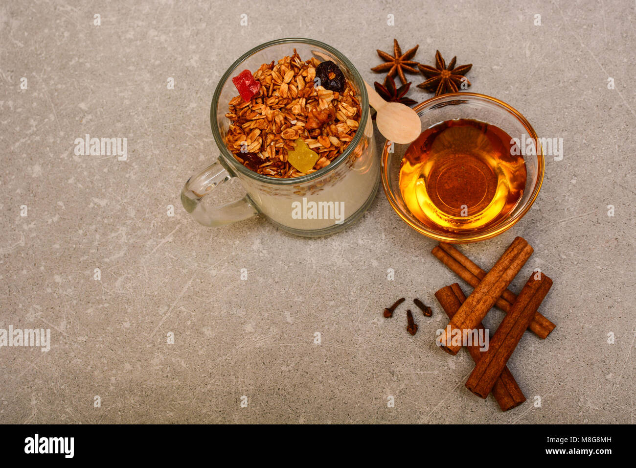 Müsli mit getrockneten Beeren, kandierten Früchten, Nüssen, Joghurt, goldenem Honig, Anis, Zimtstangen und Nelken auf steinernem Hintergrund. Stockfoto