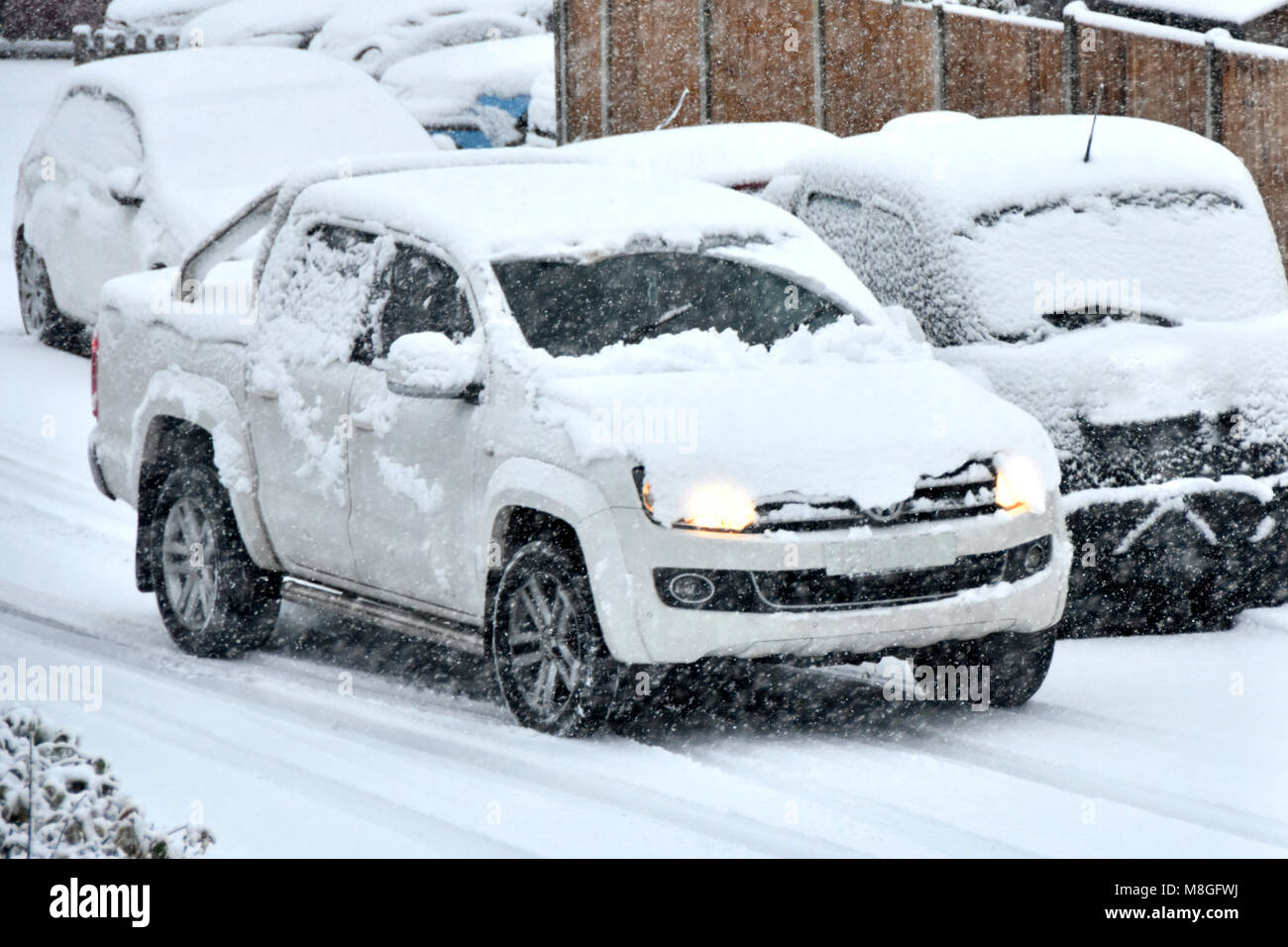 Weißes Auto im Schnee mit Scheinwerfern auf der Fahrt vorbei an geparkten Fahrzeugen mit Winter Schnee auf schneebedeckter Straße Brentwood Essex England UK abgedeckt Stockfoto