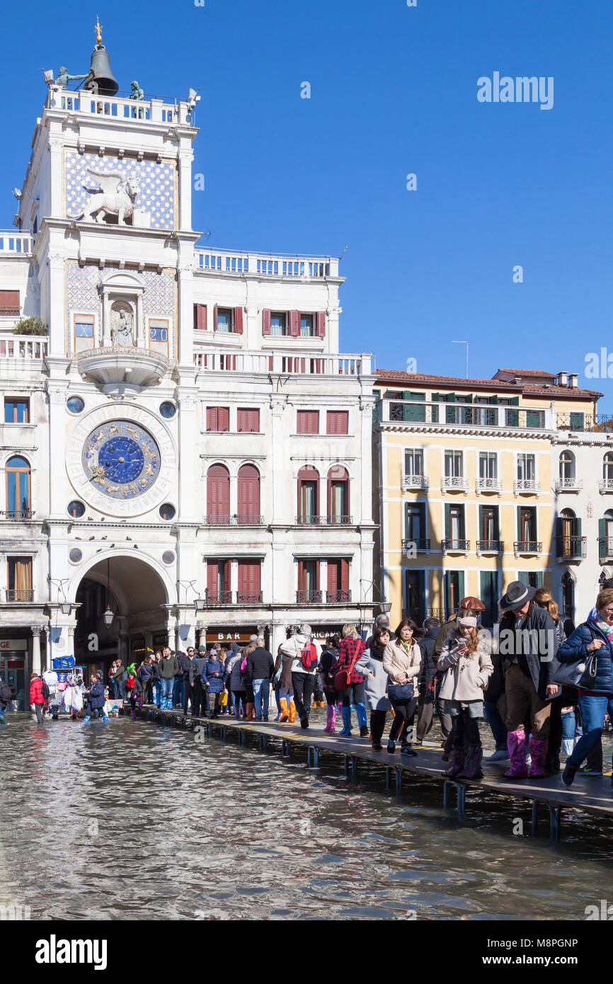 Touristen zu Fuß auf passerelles Vor dem Uhrturm durch eine überflutete Markusplatz, Piazza San Marco, Venedig, Italien während der Acqua Alta hig Stockfoto