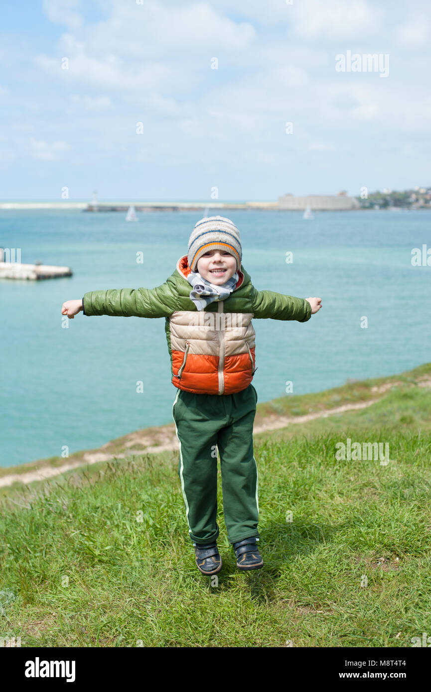 Lustige kleine Jungen in warme Jacke, Schal und Hut springt am grünen Hang gegen den Hintergrund von Meer Hafen mit Segelbooten im kalten Frühling Stockfoto