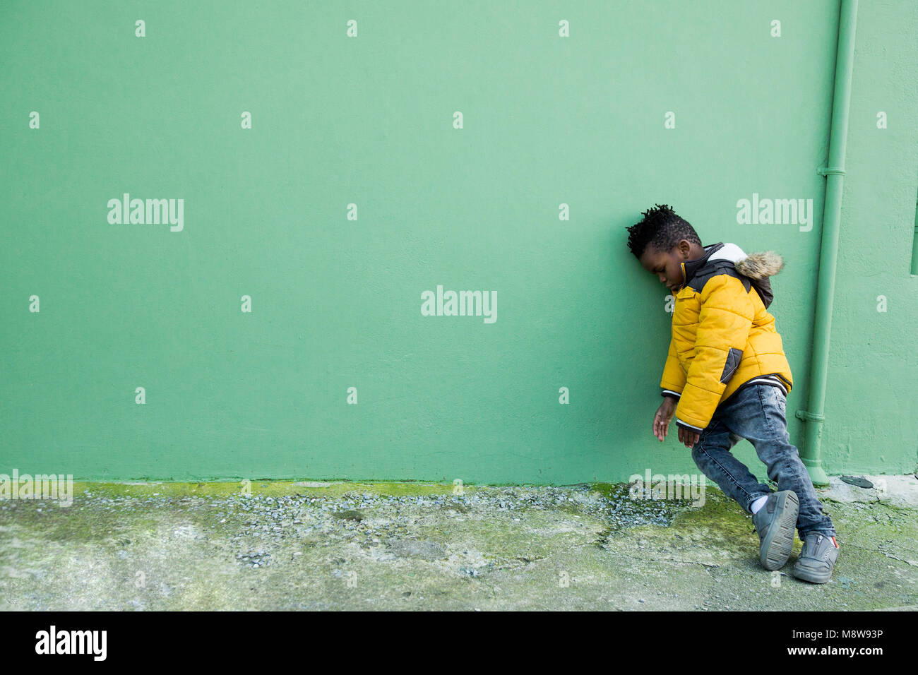 Müde und verärgert kleiner Junge im gelben Mantel lehnte sich auf einer grünen Wand auf der Straße Stockfoto