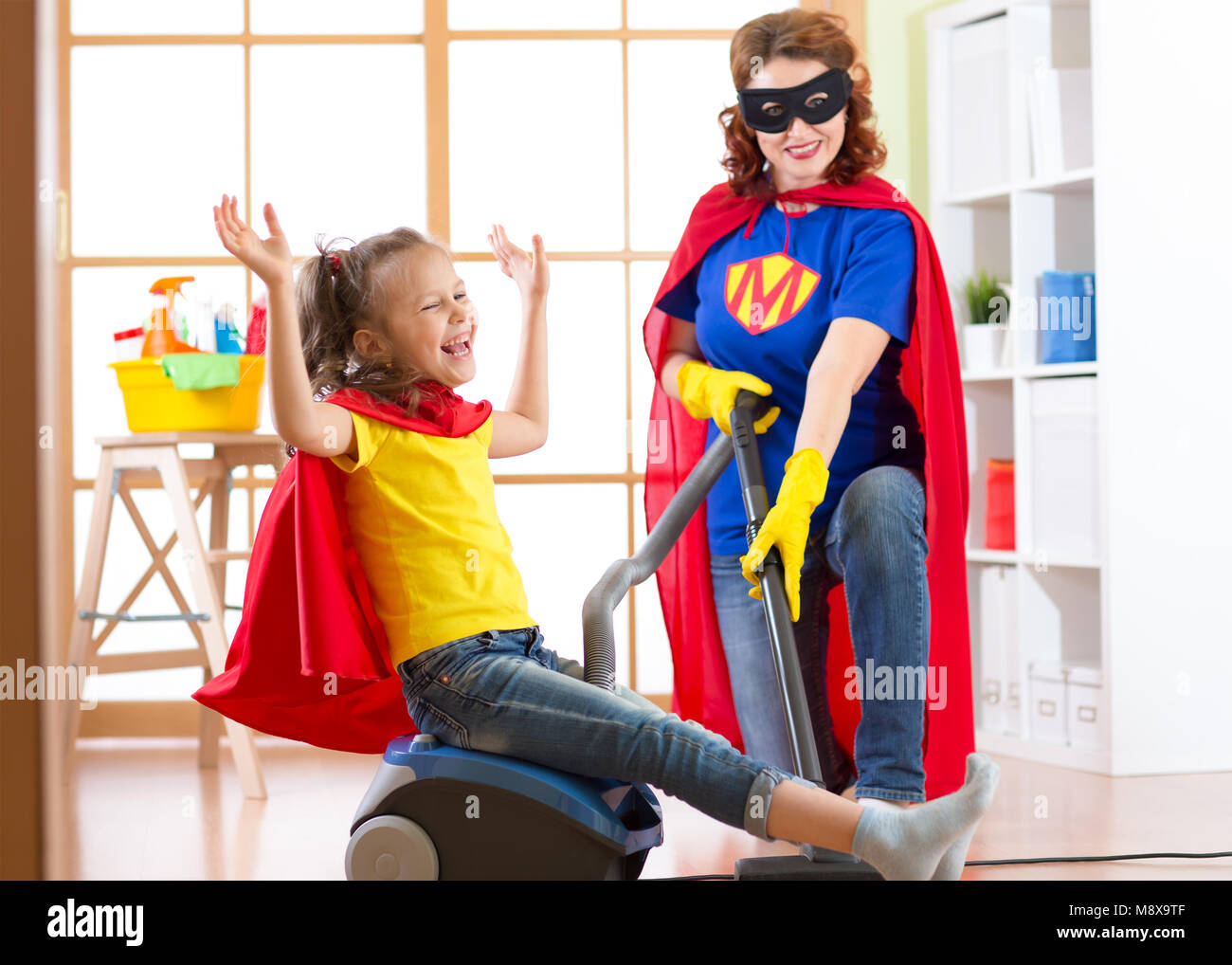 Kind und Mama verkleidet als Superhelden mit Staubsauger im Zimmer. Familie - Frau und Kind Tochter haben viel Spaß während der Bodenreinigung. Stockfoto