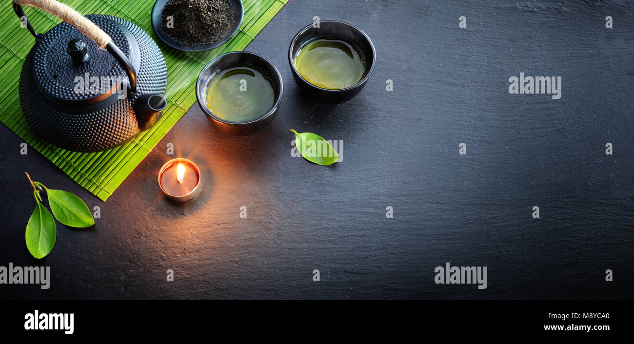 Grüner Tee in Eisen asiatische Teekanne mit Blätter und Bambus Matte auf Schwarzen Stein Stockfoto