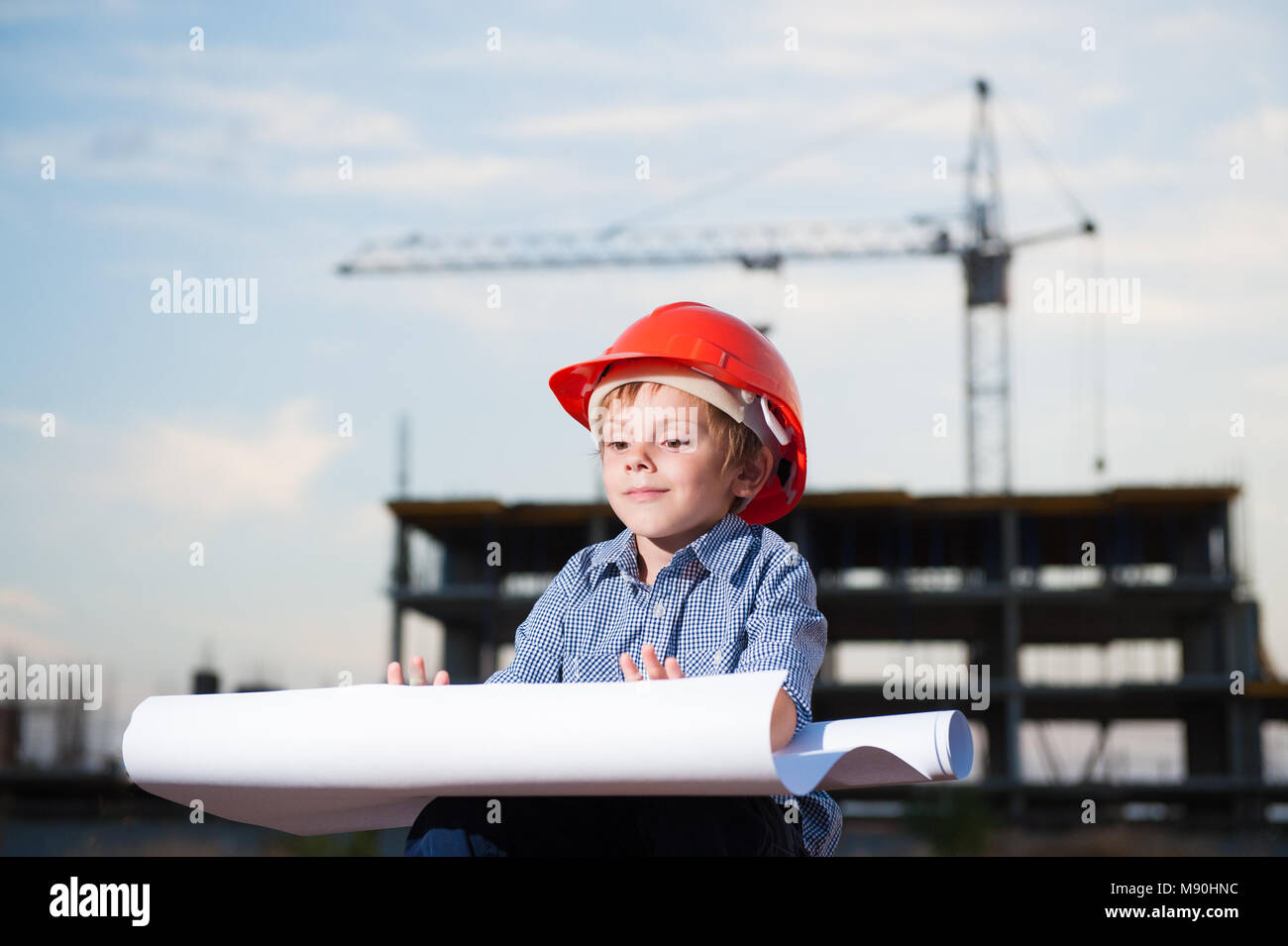 Cute little boy in orange Helm sitzt mit Papier Projekt in den Händen auf dem Hintergrund von Gebäude Bau- und Kran Stockfoto