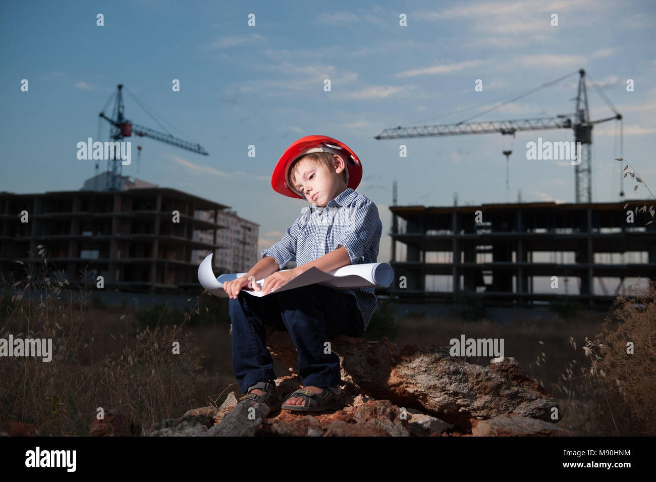 Müde kleine Junge tragen Helm löst Problem der Projekt auf der Baustelle mit Baukran im Hintergrund am Ende des Tages Stockfoto