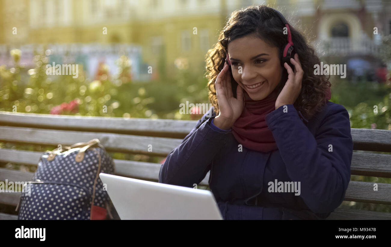 Happy girl in Kopfhörer Musik zu hören, mit freundlichen Grüßen lächelnd, sitzt auf der Bank Stockfoto