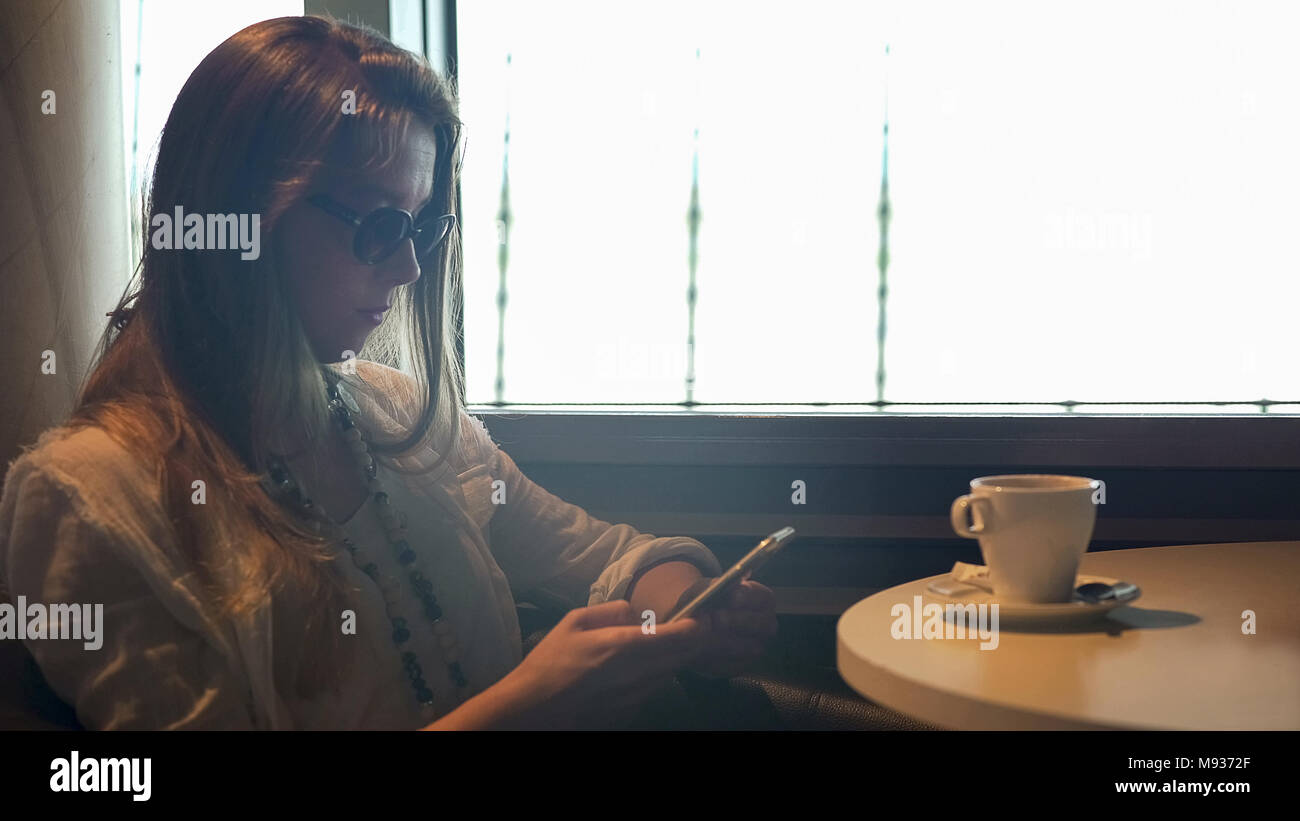 Hübsche Dame mit Sonnenbrille SMS am Telefon in ein Cafe, wartet auf Freund Stockfoto