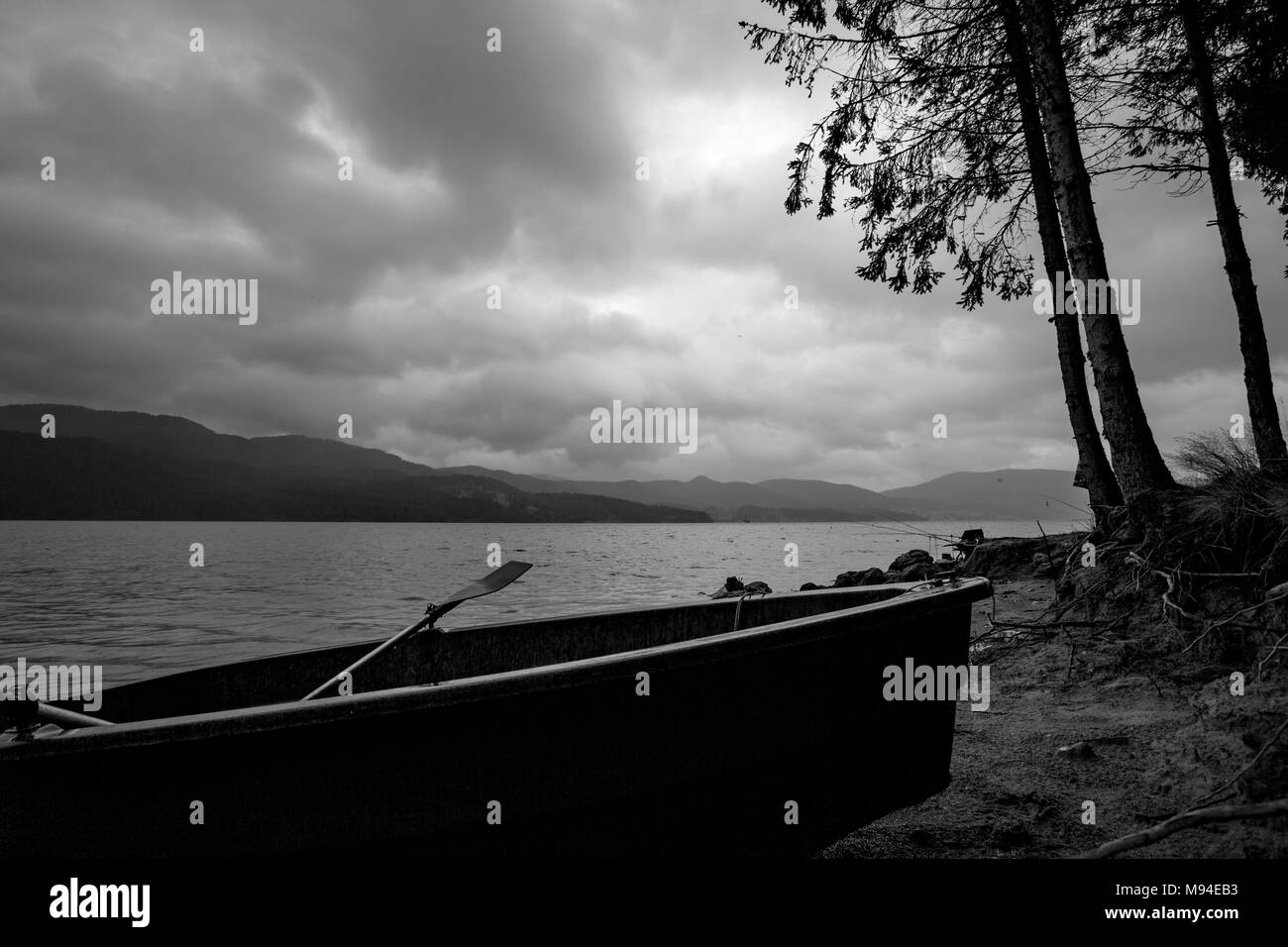 Kleines Fischerboot Silhouette am Ufer mit seitlichem Kiefern in trüben dunklen regnerischen Morgen am See Dospat, Rhodopen Gebirge, Bulgarien Stockfoto