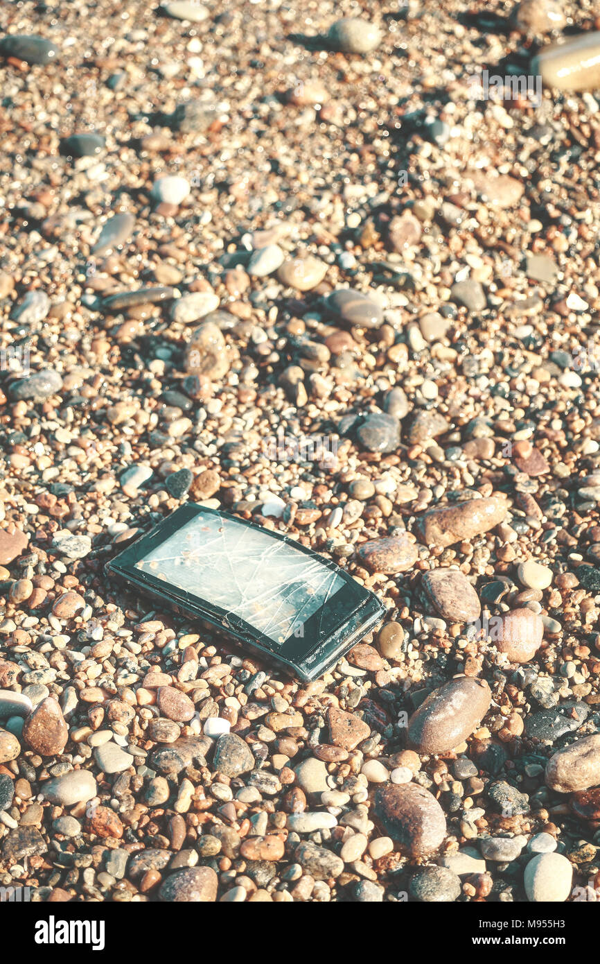 Gebrochene Handy mit gebrochenen Bildschirm auf einem steinigen Strand, selektiver Fokus, Farbe getonte Bild. Stockfoto