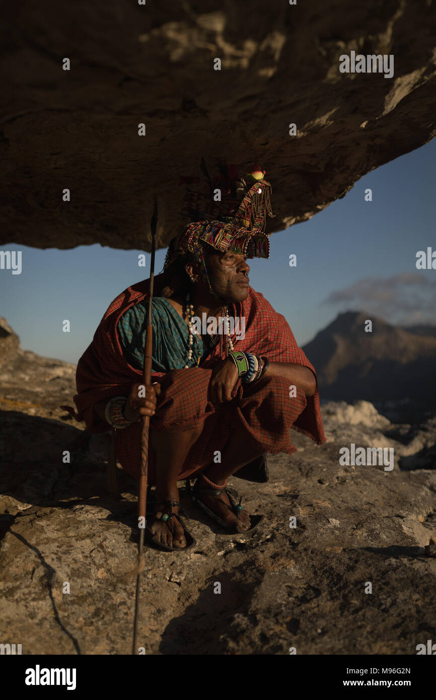 Masai Mann in traditioneller Kleidung sitzen mit Stick auf Felsen Stockfoto