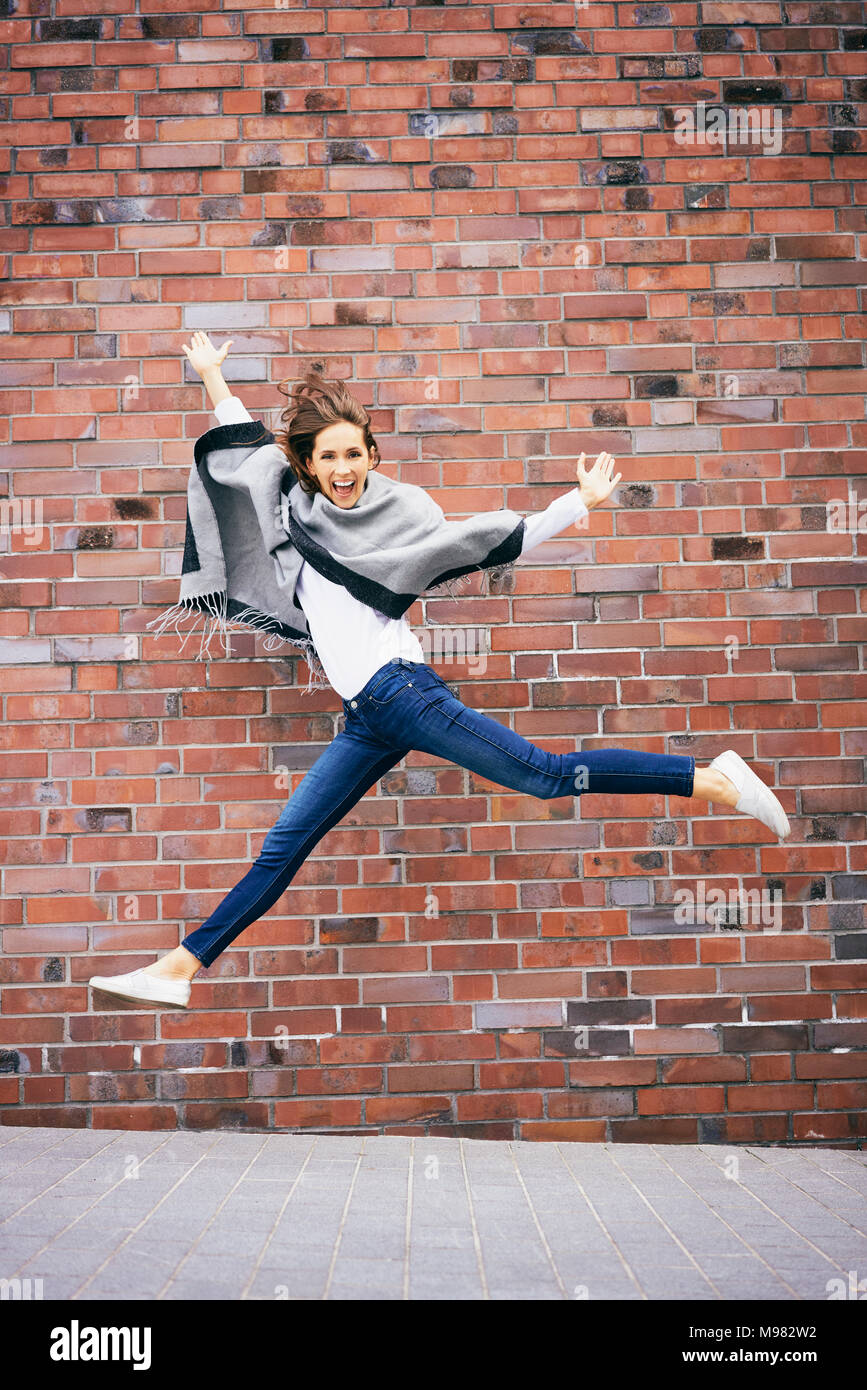 Glückliche junge Frau springt in die Luft vor Backsteinmauer Stockfoto