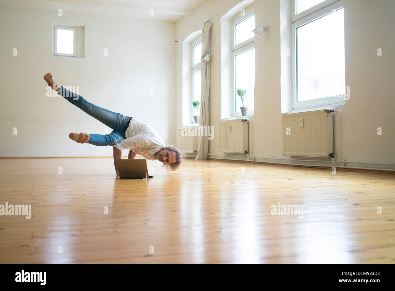 Reifer Mann macht einen Handstand auf Fußboden im leeren Raum auf der Suche nach Tablet Stockfoto