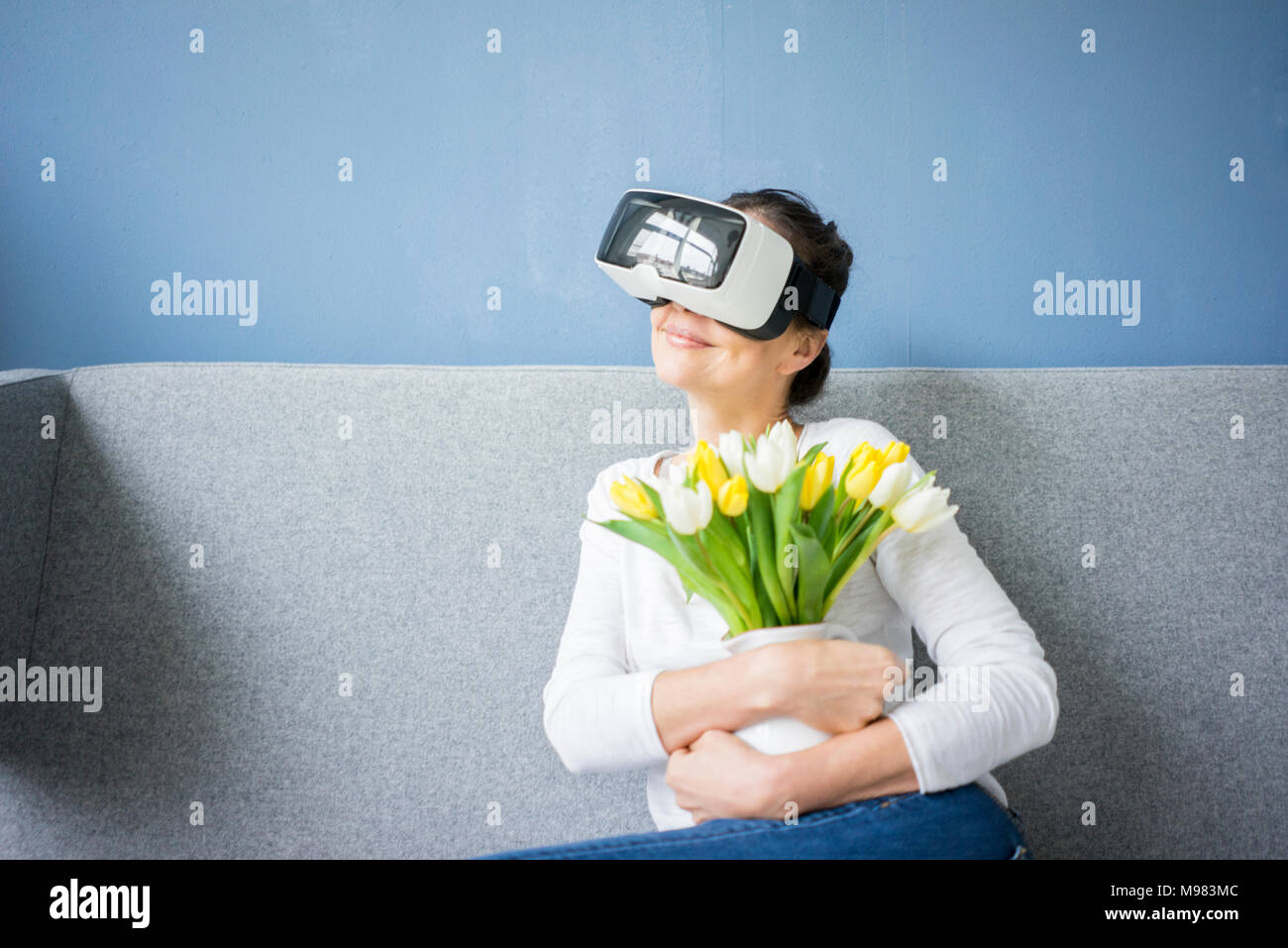 Lächelnde Frau sitzt auf der Couch tragen VR-Brille holding Bündel Tulpen Stockfoto