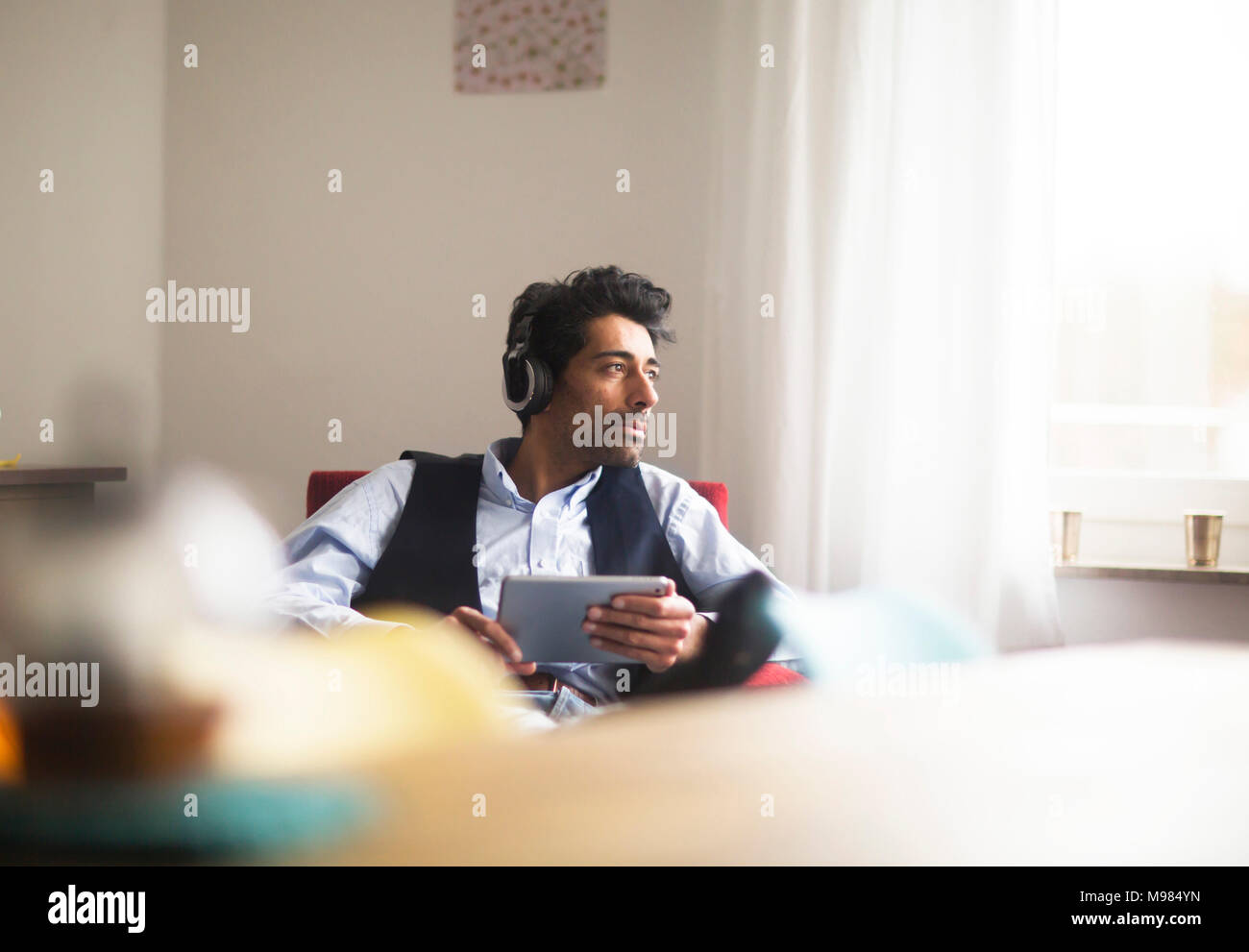 Portrait von nachdenklicher Mann mit Kopfhörer und Tablet sitzt auf einem Sessel aus Fenster Stockfoto