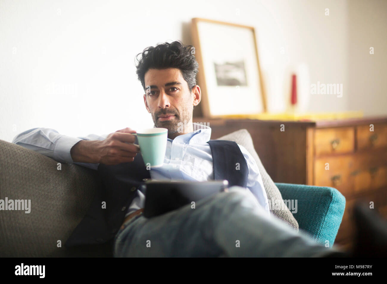 Portrait von nachdenklicher Mann mit Kaffeetasse und Tablet auf der Couch zu Hause sitzen Stockfoto