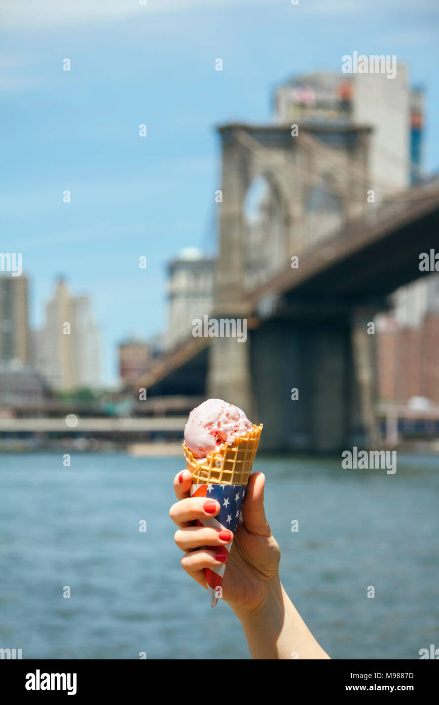 USA, New York, Brooklyn, Nahaufnahme von Frau hand mit einem Erdbeereis Kegel mit Brooklyn Bridge im Hintergrund Stockfoto