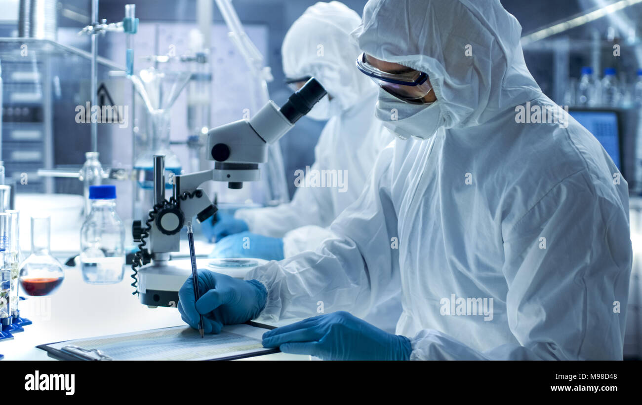 In einem sicheren Hohe Labor Wissenschaftler in einem Overall, die Durchführung einer Forschung. Chemiker mit seinem Kollegen Schreibt Experiment Ergebnisse. Stockfoto