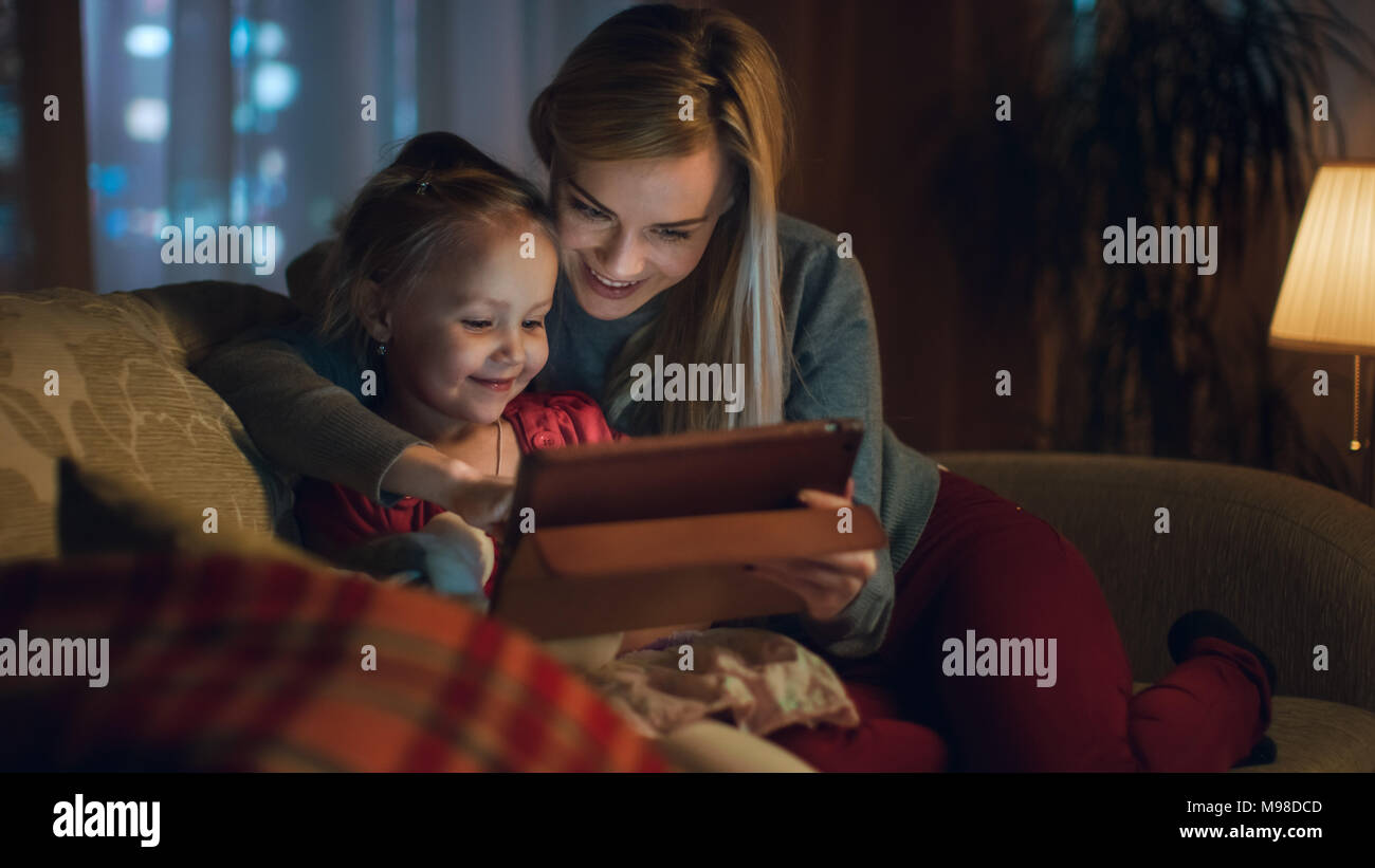 Schönen Mutter und ihre kleine Tochter auf einem Sofa im Wohnzimmer sitzen, verwenden Sie Tablet Computer. Es ist Abend, Zimmer ist gemütlich und warm. Stockfoto