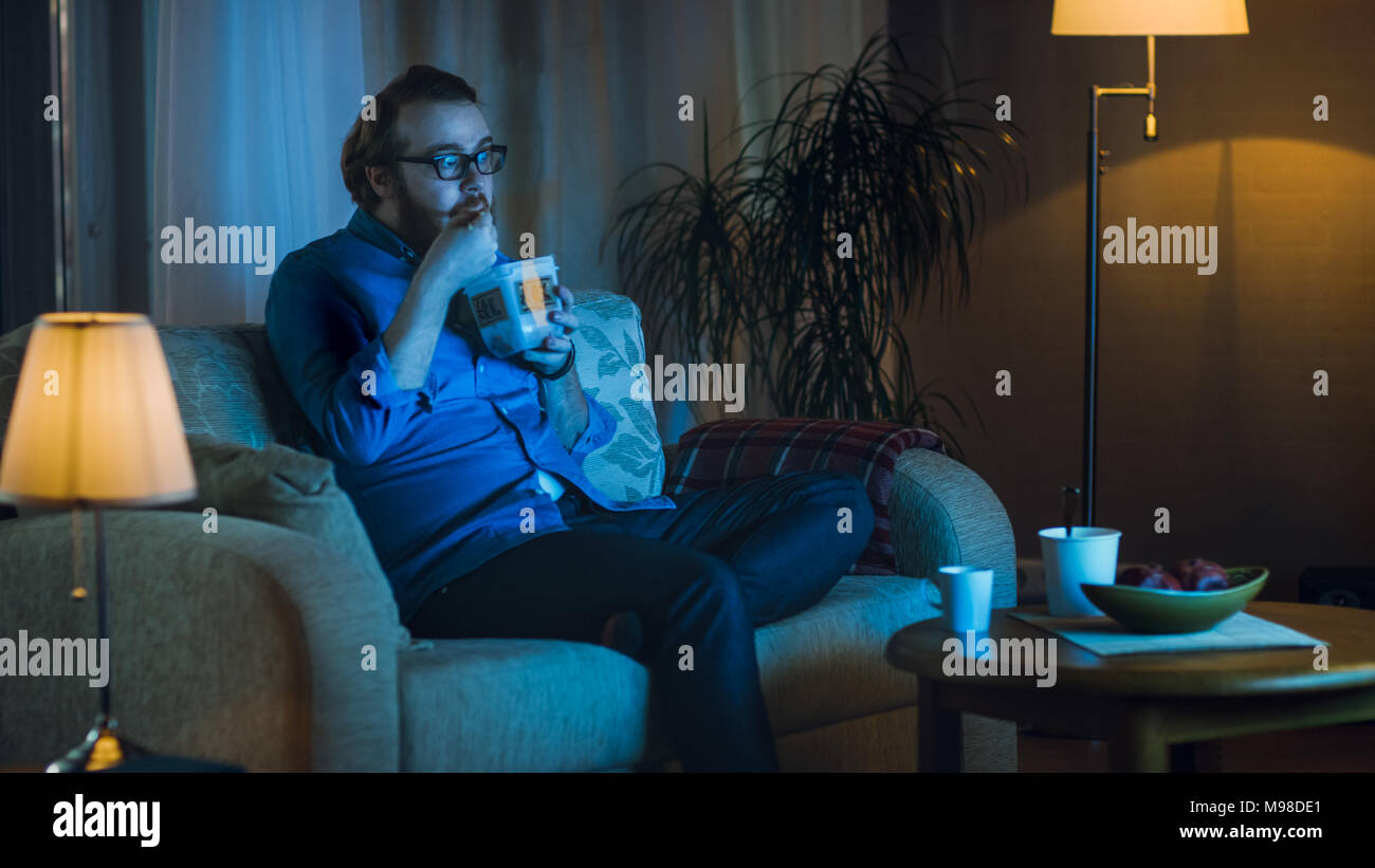 Am Abend Mann sitzt auf einer Couch in seinem Wohnzimmer. Er Essen asiatische Küche das Essen Container und Fernsehen zeigen. Stockfoto