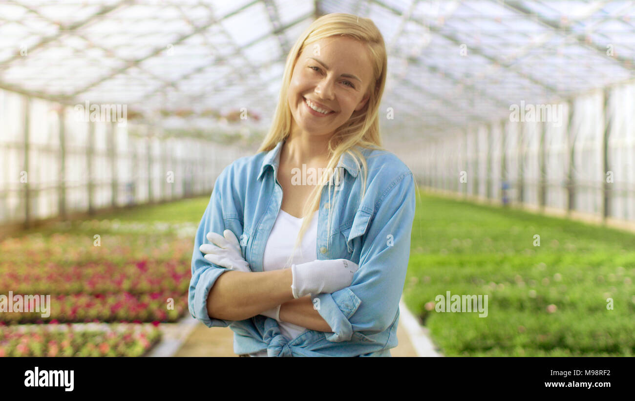 An einem sonnigen Tag schöne Blonde Gärtner steht lächelnd in einem Gewächshaus voller bunter Blumen. Stockfoto