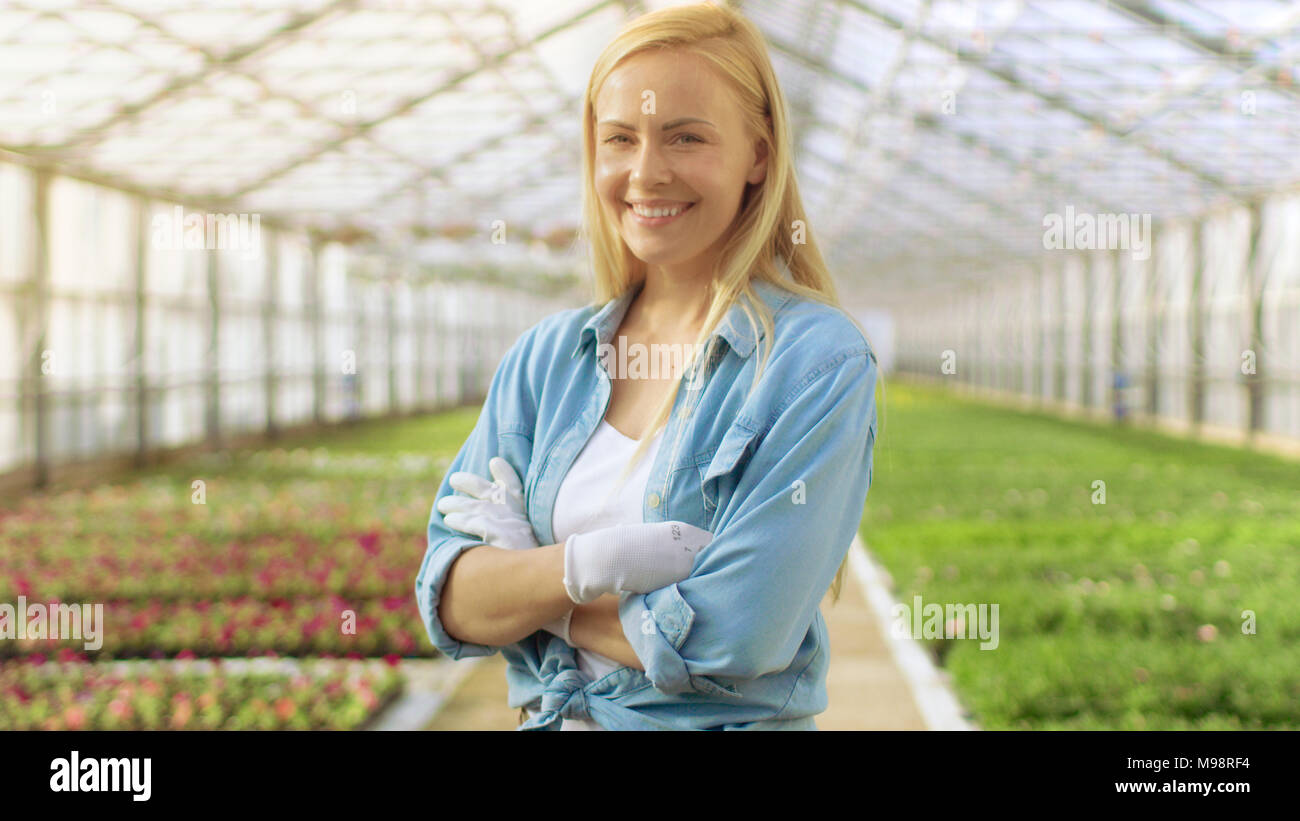 An einem sonnigen Tag schöne Blonde Gärtner steht lächelnd in einem Gewächshaus voller bunter Blumen. Stockfoto