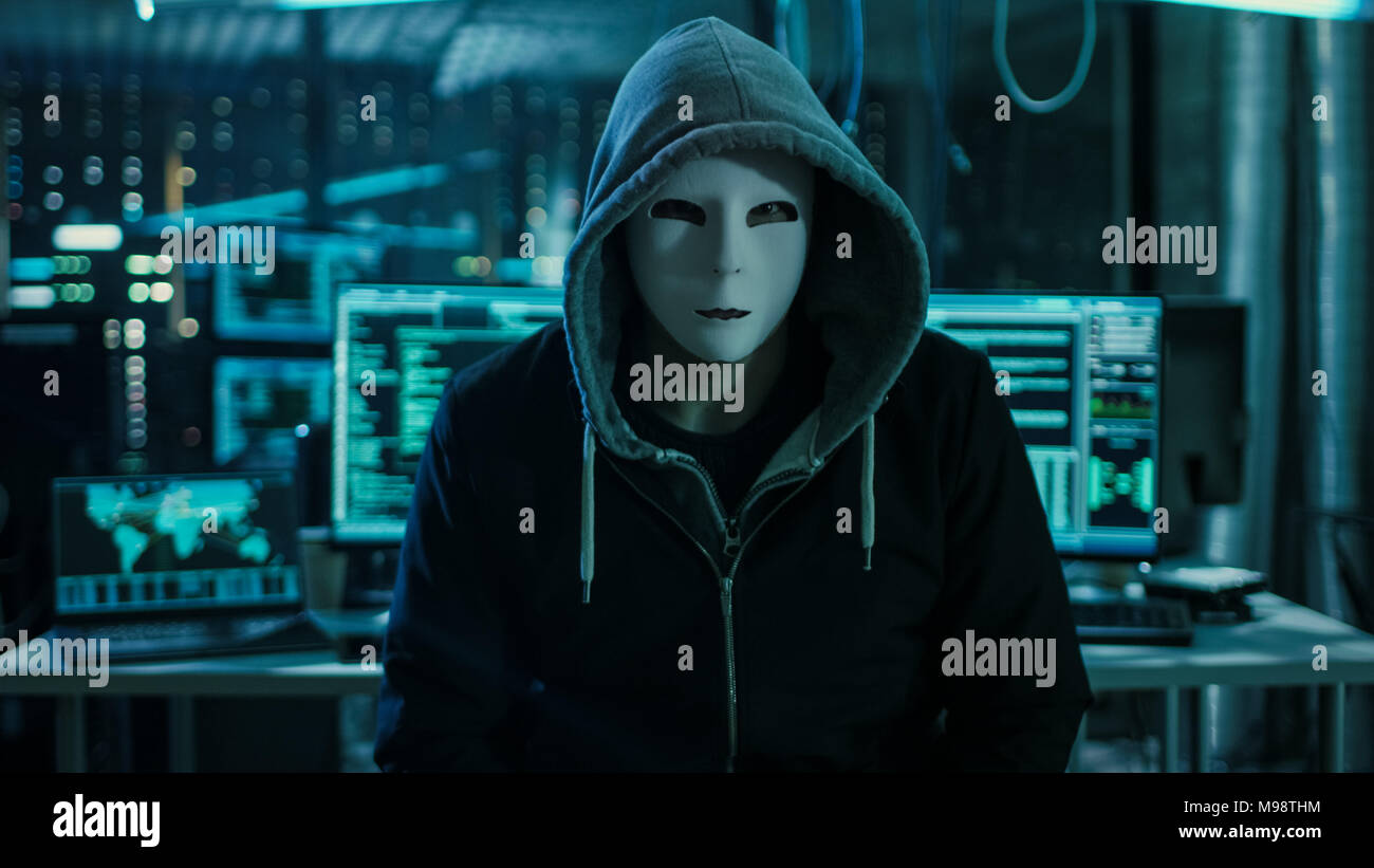 Gefährliche international gesuchten Hacker mit bedeckt Gesicht in die Kamera schaut. Im Hintergrund seine Op mit mehreren Displays Stockfoto
