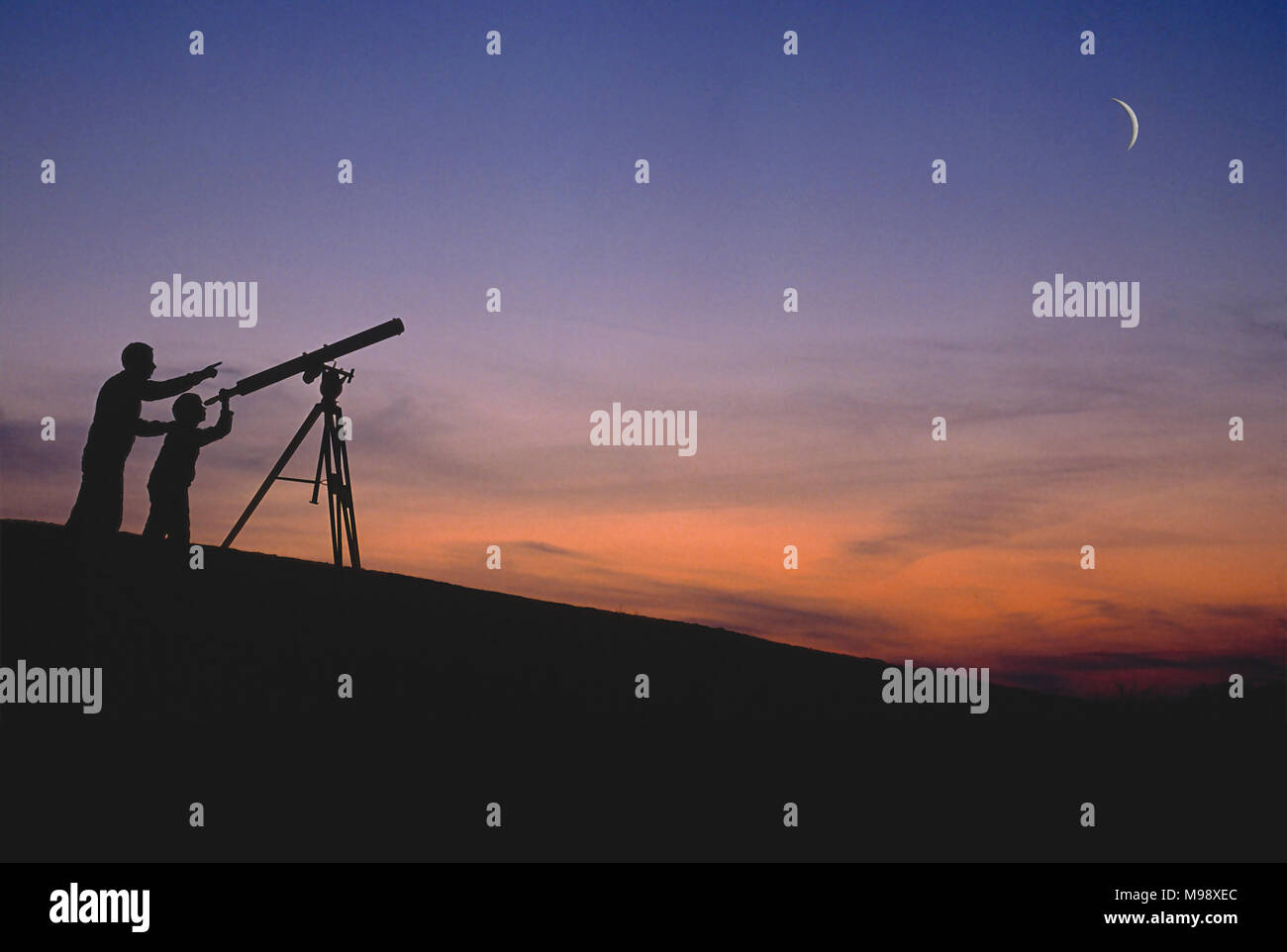 Vater unterrichtet seinen Sohn über Astronomie, Weltraum und das Universum, während sie auf einem Hügel stehen, während sie bei Sonnenuntergang den Mondsichel durch ein großes Teleskop betrachten Stockfoto