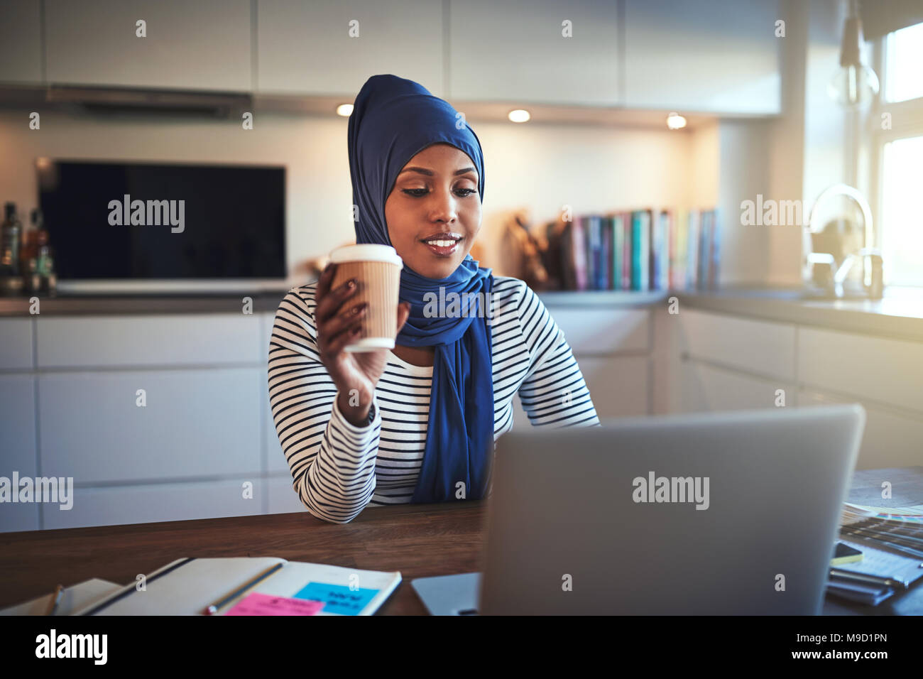 Junge arabische weibliche Unternehmer trägt ein Kopftuch, die an einem Notebook arbeitet und trinken einen Kaffee, während Sie an einem Tisch sitzen in ihrer Küche Stockfoto