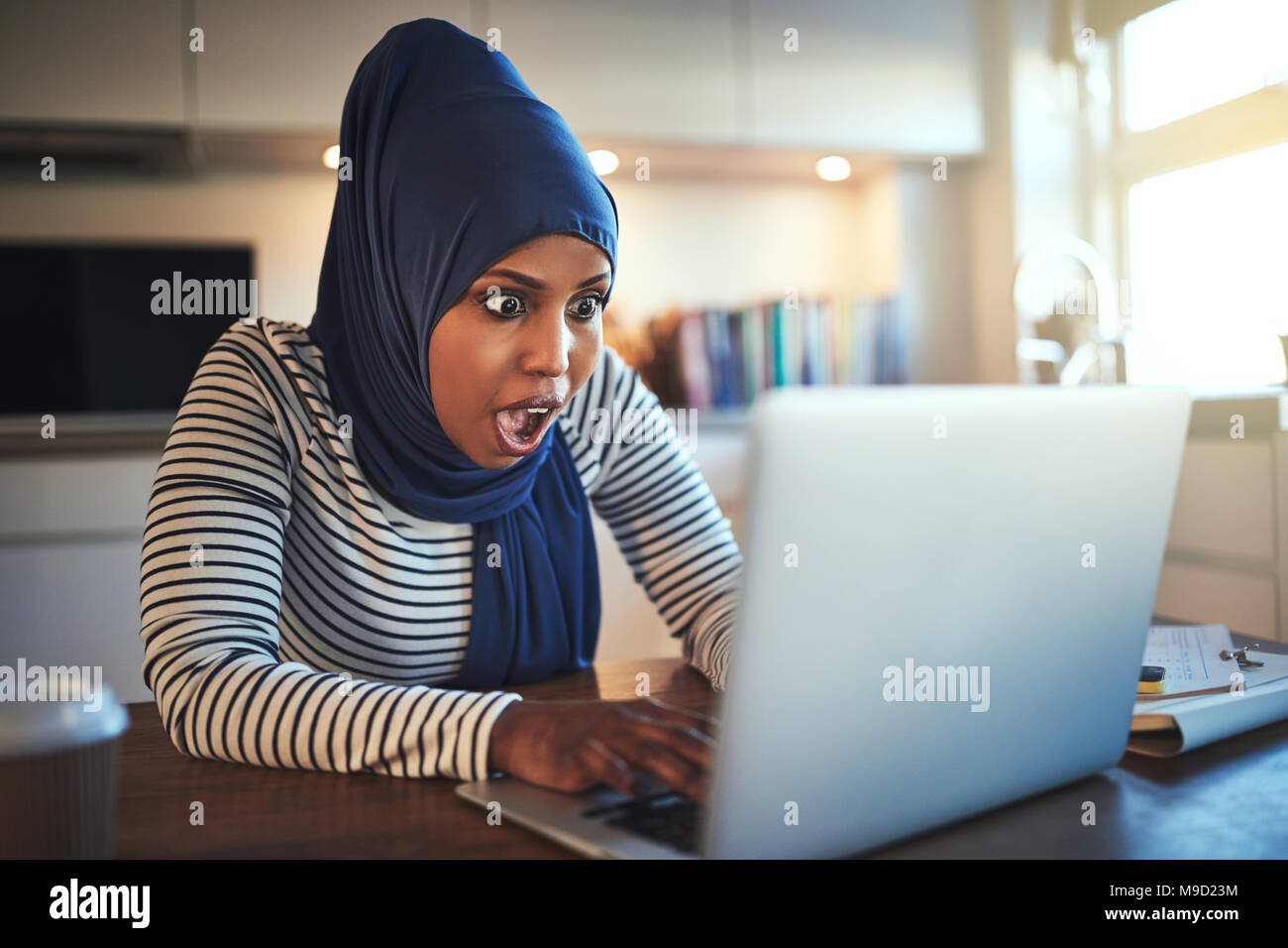 Junge arabische weibliche Unternehmer Tragen eines hijab geschockt, während an ihrem Küchentisch sitzen mit einem Laptop Stockfoto