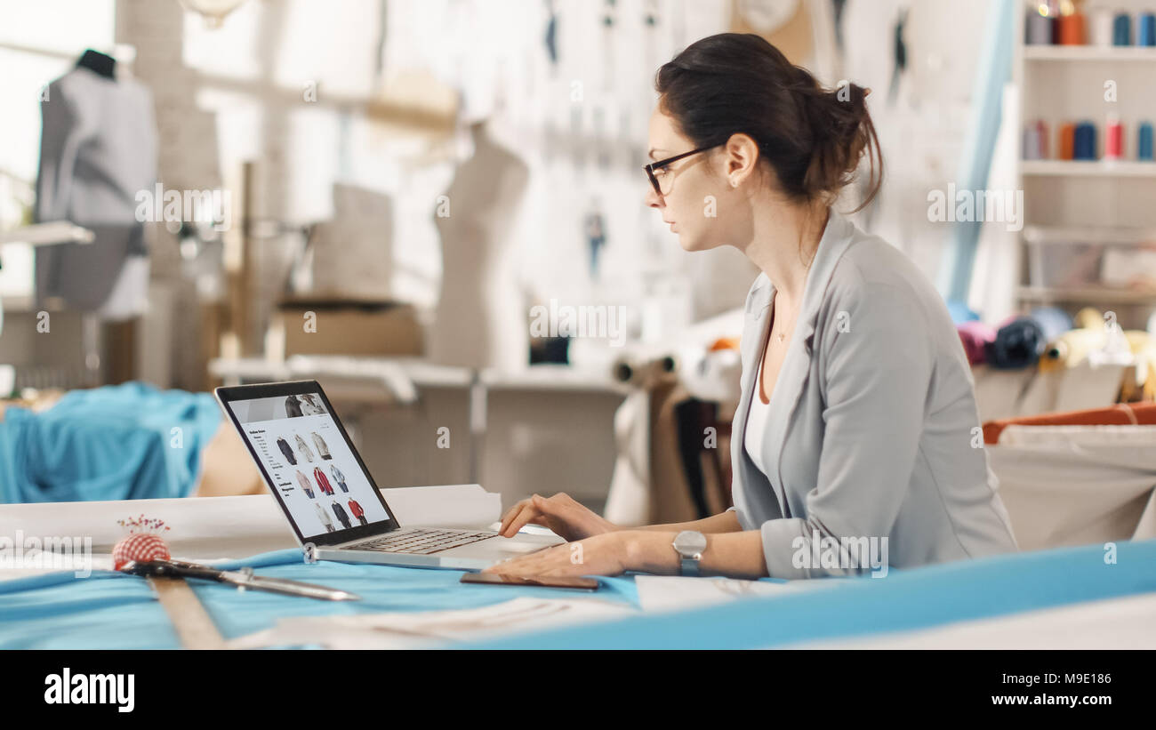 Schönen Frau, Mode, Entwerfer, Sitzen an Ihrem Schreibtisch auf Skizzen auf einem Laptop, Notizen. Ihr Studio ist sonnig, farbenfrohe Stoffe Stockfoto
