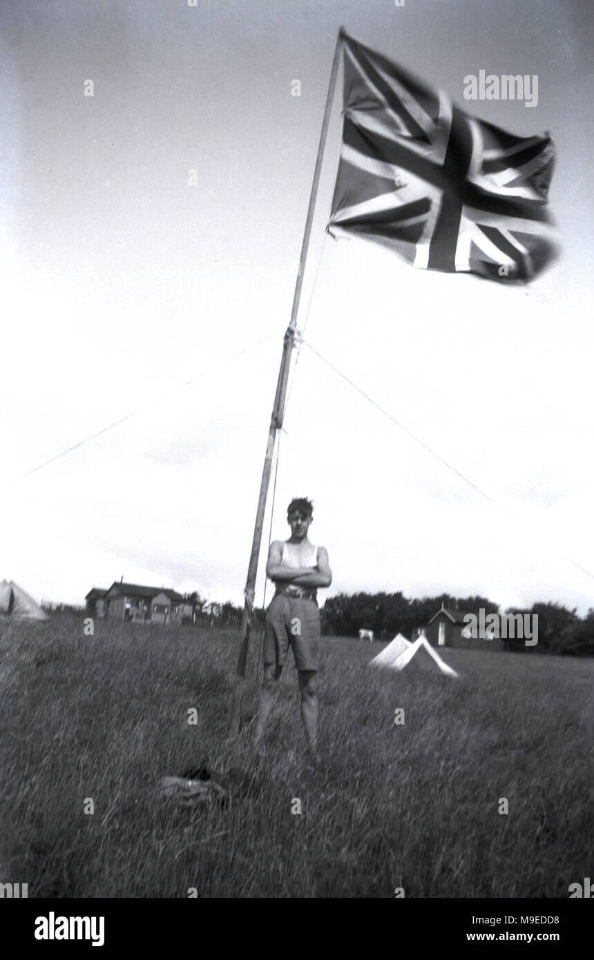 1934, historische, ein junger britischer Scout in einer Wiese zu einem Scout Camp in Dublin, Irland, steht poudly Neben hohen Fahnenmast mit einem Union Jack Flagge/ Stockfoto