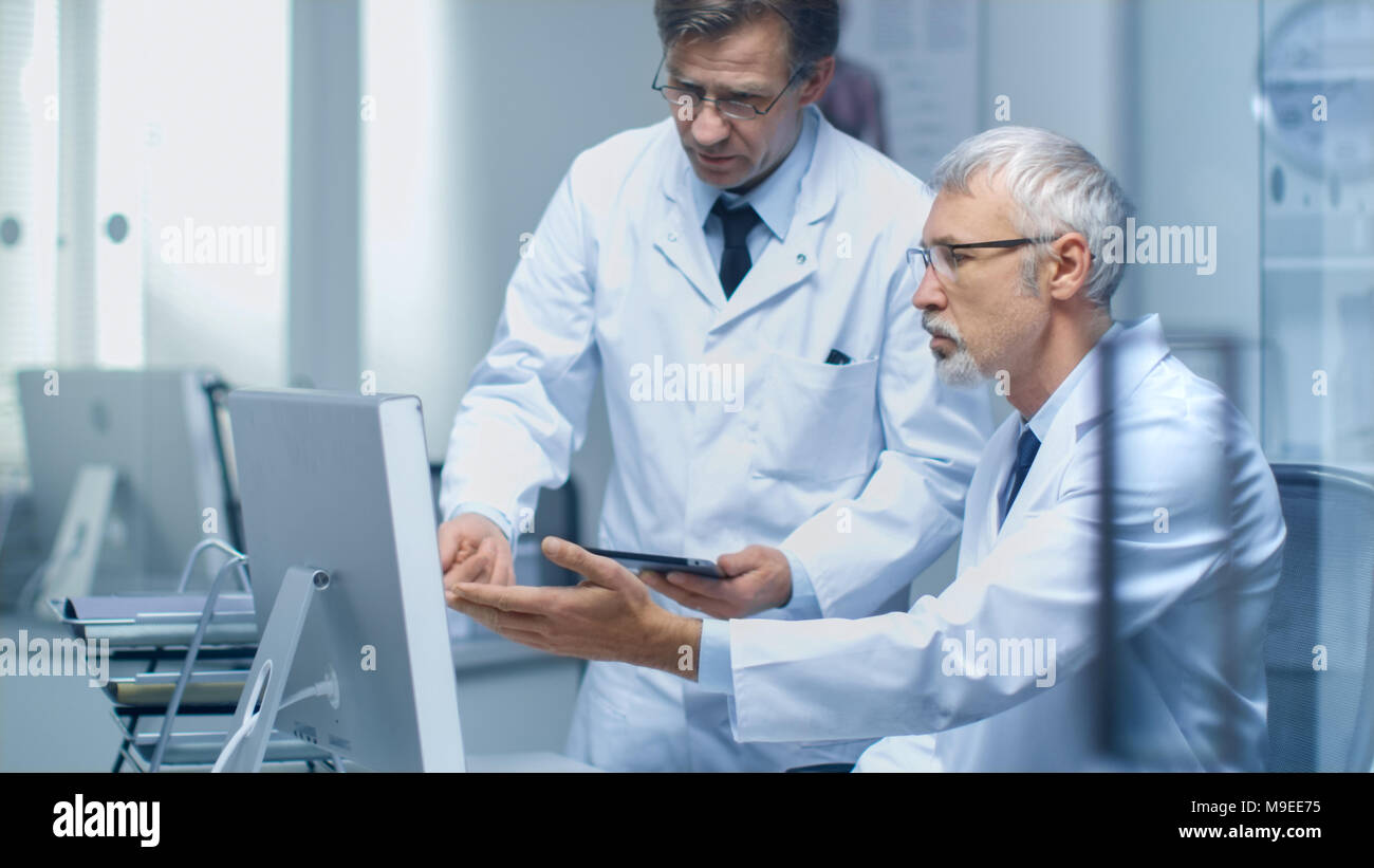 Zwei leitenden Ärzten diskutieren Medizinische Fragen über Desktop Computer. Einer von ihnen halten Tablet Computer. Büro ist modern und hell. Stockfoto