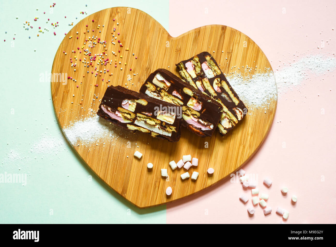 Hausgemachte Schokolade Rocky Road Kuchen in Herzform Holzbrett mit Dekorationen auf einem rosa/grünen Hintergrund Stockfoto